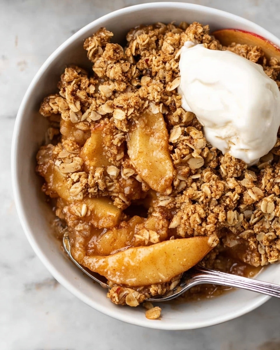A white dish with two handles holds a warm apple crumble dessert. The bottom layer shows soft, thin apple slices partially covered by a thick, golden-brown oat crumble topping that looks crunchy and textured with visible oats. On top, there are two round scoops of vanilla ice cream, smooth and creamy, slightly melting into the crumble. A spoon with a white handle and decorative silver detail rests inside the dish, scooping some of the crumble. The background is a white marbled surface with some scattered oats in a small metal measuring cup and an apple slice partially visible nearby. photo taken with an iphone --ar 4:5 --v 7
