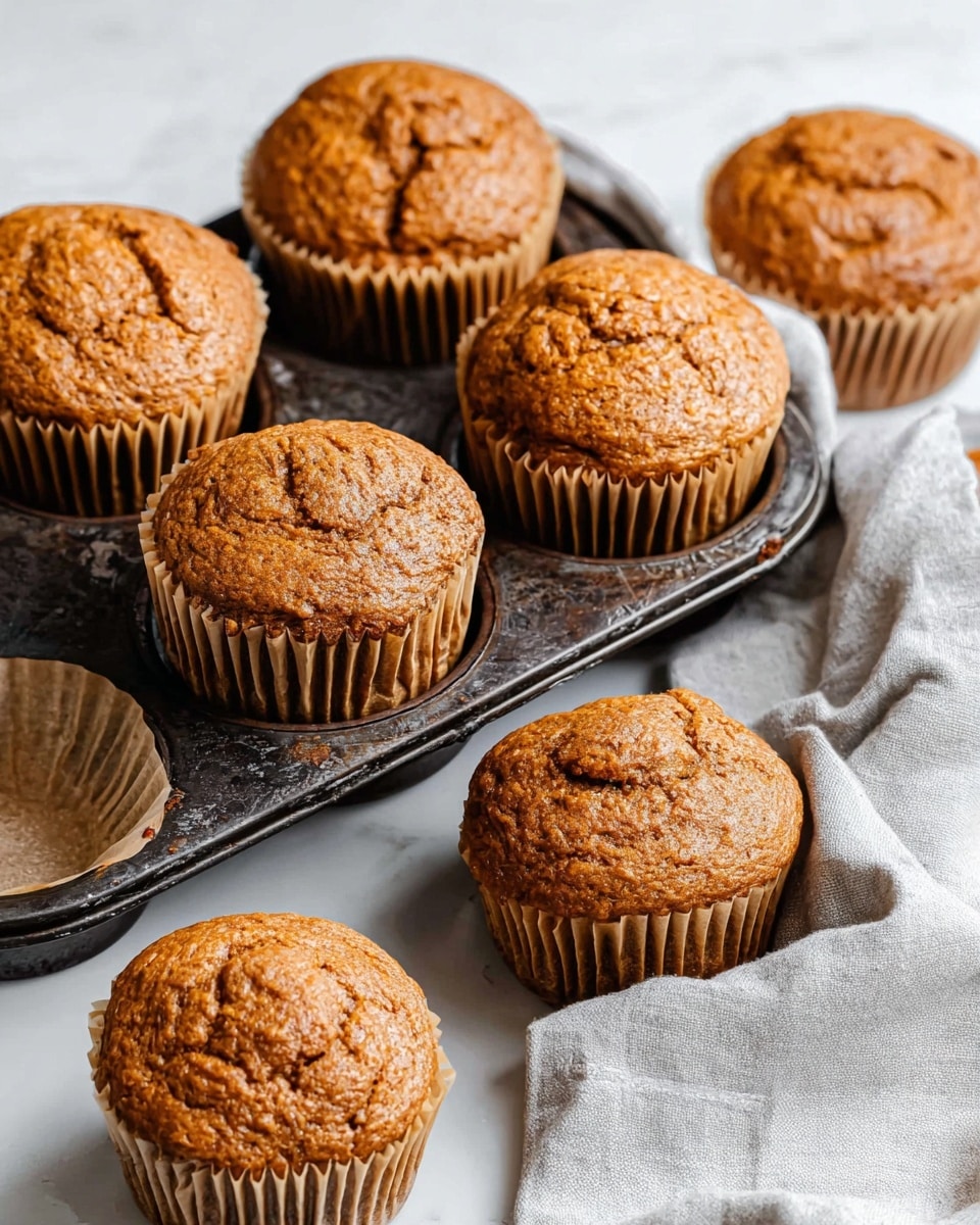 The image shows seven warm muffins, some placed inside a dark metal muffin tray and some outside on a white marbled surface. The muffins have a rough, cracked top texture with a golden brown color, each sitting in light brown paper wrappers with ridged sides. The muffin tray looks old and worn, adding a rustic feel to the scene. A light gray cloth is slightly visible on the lower right side, adding softness to the image. The lighting highlights the muffins’ slightly shiny texture, making them look fresh and inviting. Photo taken with an iphone --ar 4:5 --v 7