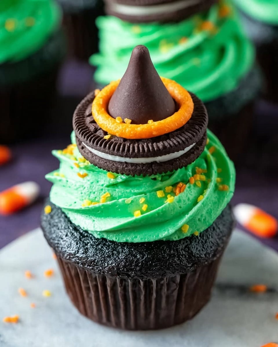 A dark chocolate cupcake with three main layers sits on a white marbled surface. The bottom layer is the dark, rich cupcake with a slight shiny texture. On top of that is a thick swirl of bright green frosting with a smooth, creamy look. Above the green frosting is a black Oreo cookie placed flat, acting like a thin layer. The Oreo is topped with a small ring of orange frosting decorated with small yellow and orange sprinkles. At the very top, there is a pointed dark chocolate kiss candy. The background includes blurred cupcakes and some scattered orange and yellow sprinkles. photo taken with an iphone --ar 4:5 --v 7