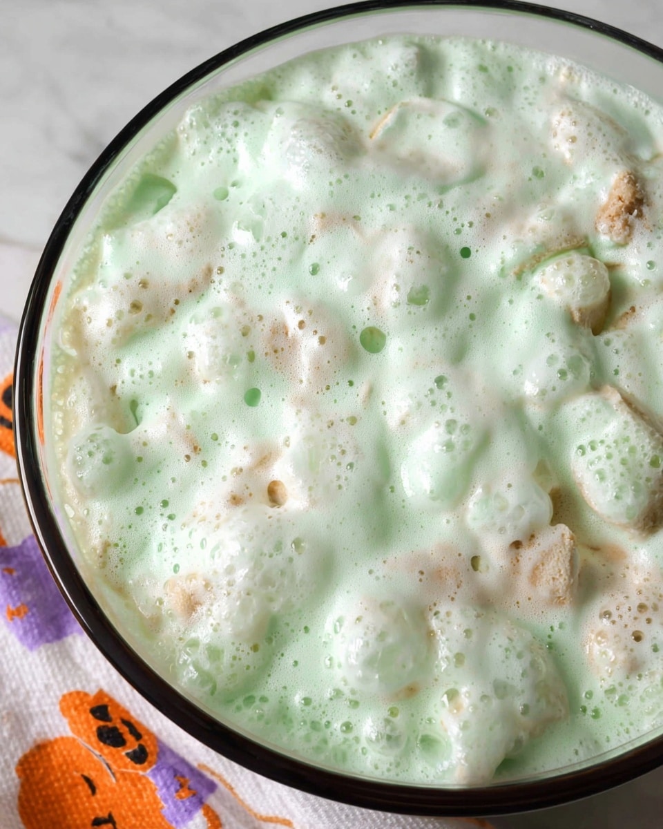 This image shows a close-up view of a glass bowl filled with a creamy dessert made of light green and white layers. The top layer is a frothy, whipped texture with soft, airy white and green bubbles covering most of the surface. Below the foam, bits of light brown and beige cake pieces can be seen, slightly soaked and soft, mixed into the creamy layers. The glass bowl has a thin black rim and sits on a white marbled surface with a white cloth that has orange and purple Halloween-themed prints partially visible at the bottom left corner. photo taken with an iphone --ar 4:5 --v 7