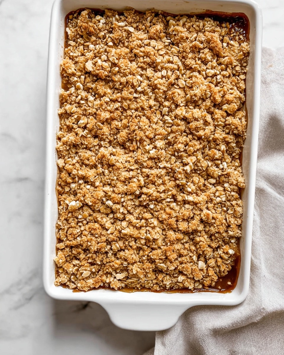 The image shows a white rectangular baking dish filled with a dessert that has two visible layers. The top layer is a rough, crumbly texture with golden brown clusters featuring oats and small pieces of nuts, covering the entire surface evenly. Just beneath the crumbly layer, a darker, glossy layer is partially visible near the edge, suggesting a soft fruit filling. The dish is placed on a white marbled surface with a light gray cloth partially visible around the bottom right corner. photo taken with an iphone --ar 4:5 --v 7