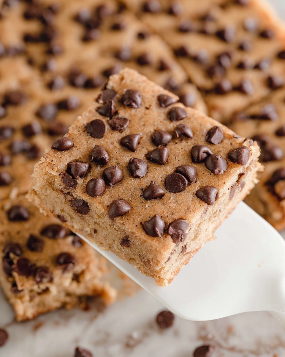A close-up view of a single square piece of a baked dessert with a light golden-brown color, dotted generously with dark brown chocolate chips all over its surface. The square piece shows a soft, slightly crumbly texture and is being lifted by a white spatula. In the background, more of the same dessert can be seen, also covered in chocolate chips, sitting on a white marbled surface. The overall look is warm and inviting, highlighting the contrast between the light brown base and the dark chocolate chips. Photo taken with an iphone --ar 4:5 --v 7