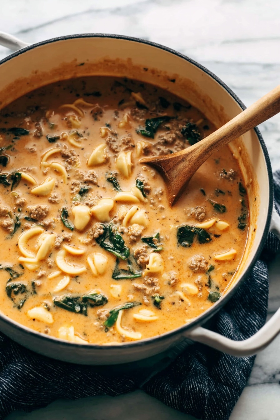 Creamy Italian Sausage Tortellini Soup, Italian sausage soup, creamy tortellini soup, comforting sausage soup recipe, easy Italian soup - A large white pot filled with a thick, creamy soup that is orange-beige in color, speckled with small pieces of ground meat, green leafy vegetables, and small curved pasta shells. A wooden spoon is resting inside the pot, partially submerged in the soup, showing its rich and smooth texture. The pot sits on a dark blue cloth on a white marbled surface, creating a warm and cozy feeling. photo taken with an iphone --ar 2:3 --v 7