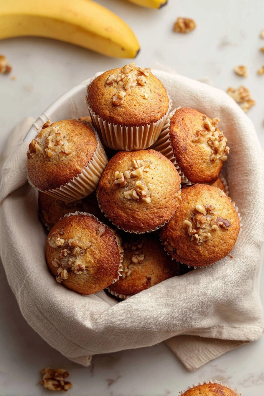 Quick & Easy Vegan Banana Muffins, vegan banana muffins, healthy banana muffin recipe, plant-based banana muffins, quick vegan breakfast muffins - A white bowl lined with a soft beige cloth holds a pile of golden brown muffins with slightly domed tops. Each muffin is in light beige paper cups and sprinkled with small pieces of walnuts on the top, giving a rough texture. Around the bowl on a white marbled surface, there are a few loose walnuts and bananas visible, adding natural color contrasts. The muffins appear moist with a lightly browned crust. photo taken with an iphone --ar 2:3 --v 7