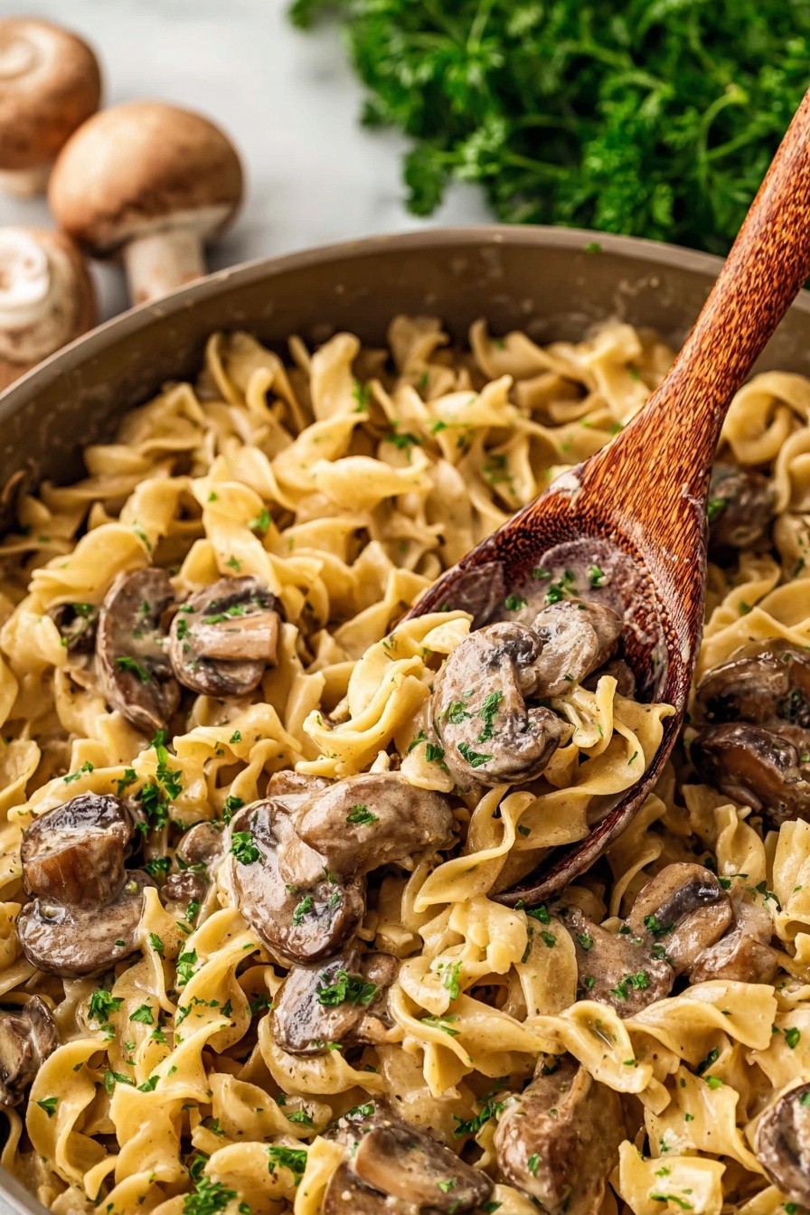 Old Fashioned Beef Stroganoff, beef stroganoff recipe, comfort food recipes, classic beef stroganoff, easy beef stroganoff - The image shows a close-up of a pan filled with a creamy noodle and mushroom dish. There are egg noodles that are light yellow, soft, and curly, spread all over the pan as the first layer. On top of the noodles, there are sliced mushrooms with a brown color and slightly shiny surface from the sauce. Mixed with the mushrooms and noodles, there are small bits of green parsley scattered for color. A wooden spoon is scooping some of the dish from the pan. The background has some blurred green parsley and whole mushrooms placed on a white marbled surface. photo taken with an iphone --ar 2:3 --v 7