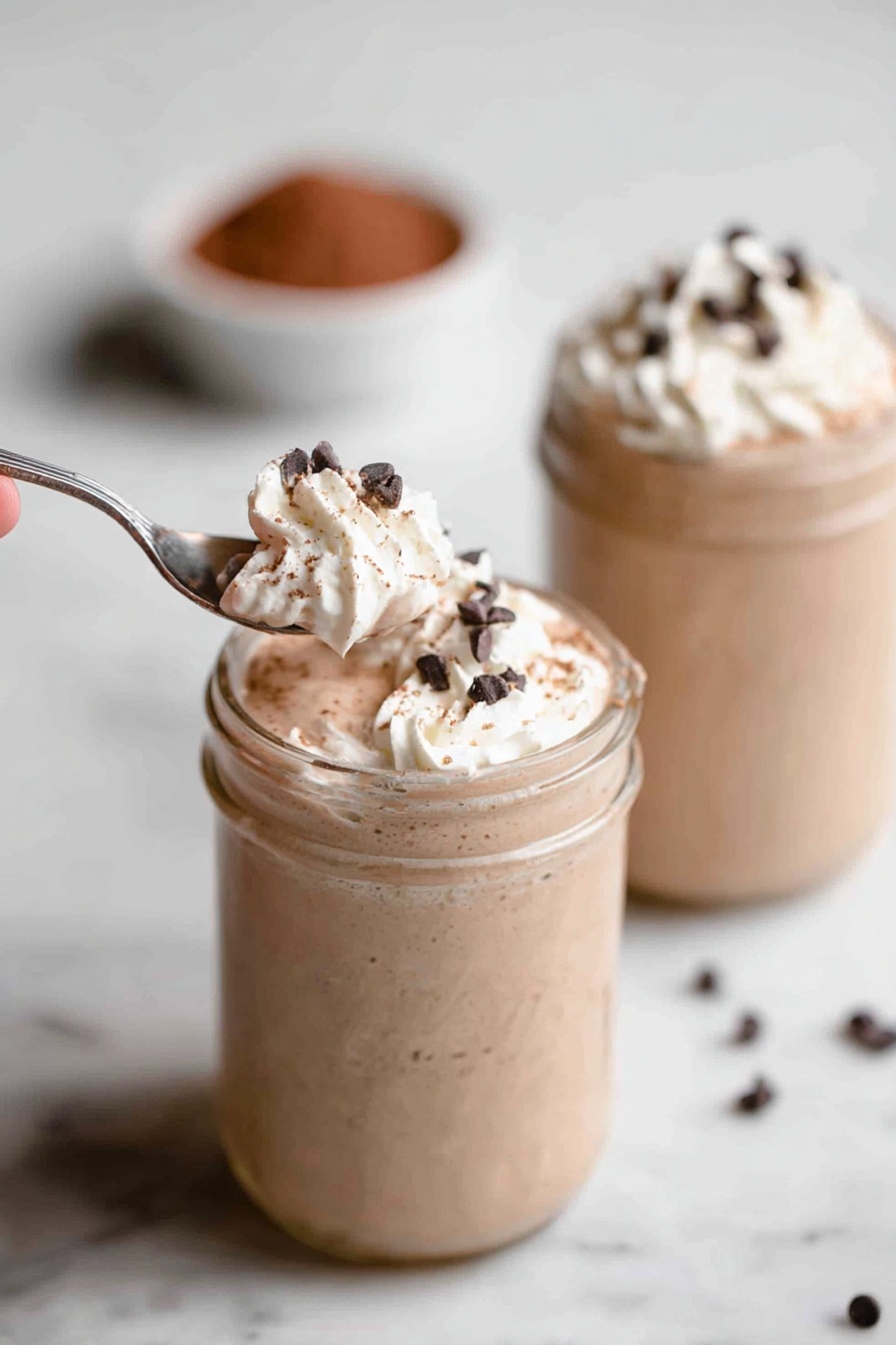 Vegan Chocolate Peanut Butter Protein Shake, healthy vegan protein shakes, plant-based chocolate peanut butter smoothie, quick vegan protein drinks, dairy-free chocolate peanut butter shake - Two glass jars sit on a white marbled surface, each filled with a creamy light brown milkshake. The milkshakes have a thick texture and are topped with a swirl of white whipped cream. Small, dark chocolate chips are sprinkled over the whipped cream. In the foreground, a silver spoon held by a woman's hand scoops a portion of whipped cream and chocolate chips from the first jar. The background jar is out of focus but shows the same whipped cream and chocolate chip topping. There is a small bowl filled with cocoa powder blurred in the background. Photo taken with an iphone --ar 2:3 --v 7