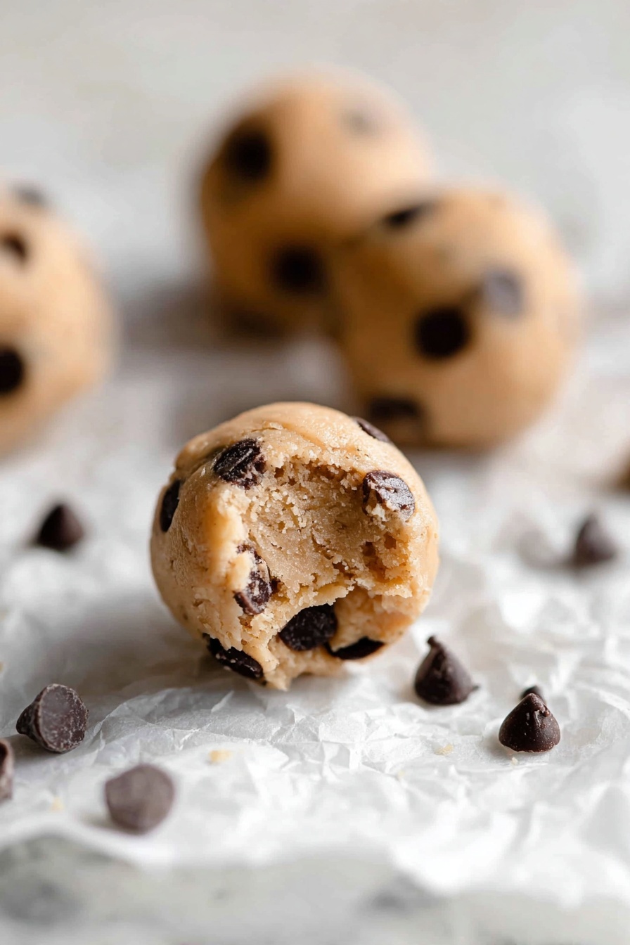 Healthy Protein Cookie Dough Bites, healthy snack recipes, no-bake protein snacks, quick healthy treats, protein powder cookie bites - The image shows small round cookie dough balls with a light tan color, filled with many dark brown chocolate chips scattered inside and on the surface. The balls rest on a crumpled white paper on a white marbled background. One dough ball in the front has a small bite taken out of it, showing a soft, slightly crumbly texture inside, while other dough balls and some loose chocolate chips are blurred in the background. The lighting is soft and natural, highlighting the texture of the dough and chocolate chips, photo taken with an iphone --ar 2:3 --v 7