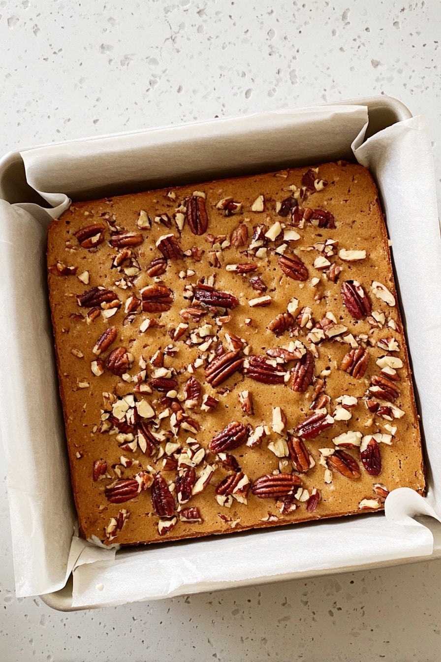 Maple Pecan Blondies, maple pecan blondies recipe, easy blondies with pecans, comforting fall desserts, homemade blondie bars - A square baked dessert with a light brown top covered in scattered pieces of pecans. It is in a white baking pan lined with parchment paper that folds over the edges. The baked item looks soft and slightly puffy, with a smooth but slightly textured surface from the nuts. The pan sits on a white marbled surface. photo taken with an iphone --ar 2:3 --v 7