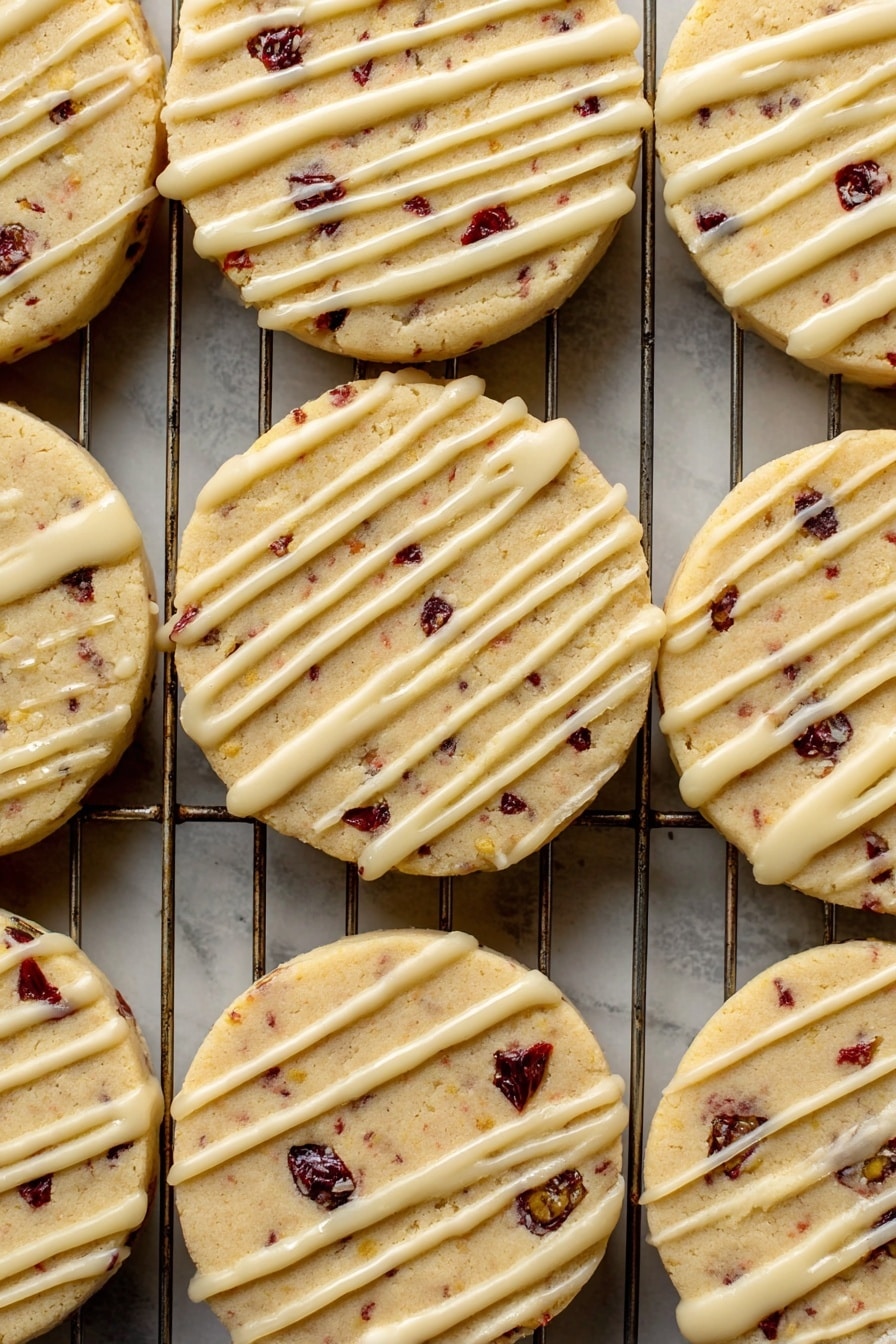 Orange Cranberry Shortbread Cookies, cranberry shortbread cookies, orange zest cookies, holiday shortbread recipes, buttery citrus cookies - A close-up view of a batch of round cookies laid out on a metal cooling rack over a white marbled surface. Each cookie has one visible layer, light golden in color with small dark red and black fruit bits inside, giving a spotted look. On top of each cookie is a thin, even drizzle of pale cream-colored icing, applied unevenly in long lines across the surface. The texture of the cookies looks soft but firm, with some slight cracks near the edges. The metal wires of the rack create a grid pattern underneath the cookies. Photo taken with an iphone --ar 2:3 --v 7
