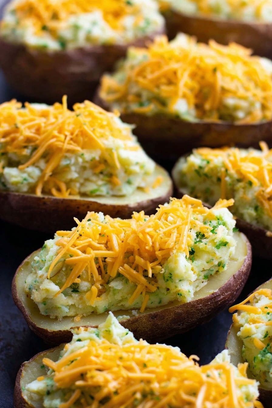 Vegan Cheesy Broccoli Twice Baked Potatoes, vegan stuffed potatoes, healthy vegan dinner, dairy-free cheesy potatoes, plant-based baked potato dish - The image shows several potato halves with the inside scooped out and filled with a creamy mixture that looks light yellow with green bits throughout, suggesting herbs or vegetables mixed in. Each stuffed potato is topped with a generous layer of shredded orange cheddar cheese. The potatoes are arranged closely together on a dark surface which contrasts with the light filling and bright cheese. The texture of the filling appears smooth but slightly chunky, and the potato skins are a dark brown color. The close-up shot highlights the layers clearly with the cheese on top, the creamy filling in the middle, and the potato skin at the bottom. Photo taken with an iphone --ar 2:3 --v 7