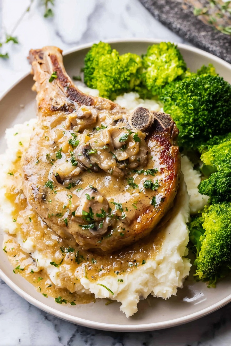Easy Smothered Pork Chops, Pork Chops with Onion Mushroom Gravy, Hearty Pork Chop Dinner, Simple Pork Chop Recipe, Comfort Food Pork Chops - A large cooked pork chop sits in the center of a white plate on a bed of creamy mashed potatoes as the bottom layer; the pork chop has a light brown color with a creamy mushroom sauce on top, dotted with pieces of mushrooms and sprinkled with small green herbs; to the right side of the plate, there are bright green steamed broccoli florets with a slightly wet texture; the white mashed potatoes have a soft, slightly lumpy texture, and both the pork chop and sauce have a glossy sheen from the sauce; the plate sits on a white marbled surface; photo taken with an iphone --ar 2:3 --v 7