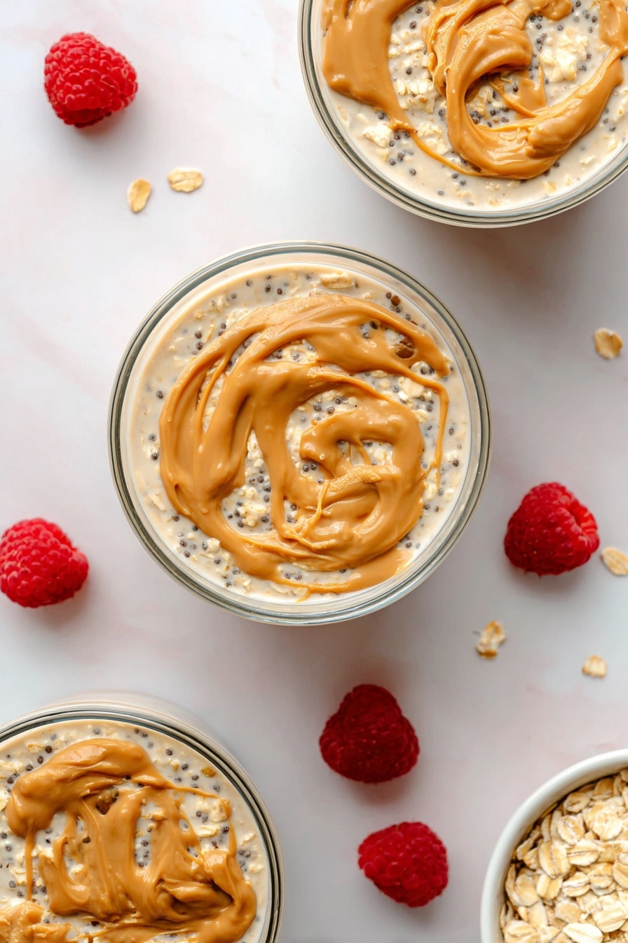 Peanut Butter Overnight Oats, healthy breakfast ideas, easy overnight oats, nutritious breakfast recipes, vegan overnight oats - The image shows three round clear glass bowls filled with layered overnight oats on a white marbled surface. Each bowl has a bottom layer of creamy light beige oats mixed with small black chia seeds, covered by a thin layer of milky liquid. The top layer is swirled with dollops of light brown peanut butter, creating smooth, curved textures on the surface. Around the bowls, scattered fresh bright red raspberries add a pop of color, along with a small white bowl filled with rolled oats placed in the bottom right corner. The photo taken with an iphone --ar 2:3 --v 7