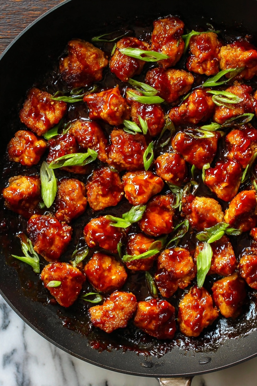 Honey Ginger Seitan with Crispy Pan-Fried Glazed Bites, vegan seitan recipe, plant-based honey ginger bites, crispy glazed vegan appetizer, savory vegan seitan dish - This image shows a black pan filled with many small pieces of golden-brown chicken covered in thick, shiny reddish-brown sauce. The pieces are spread out evenly across the pan, with some bright green slices of scallion sprinkled on top. The sauce looks sticky and glossy, covering all the chicken pieces in a rich layer. The pan's dark color contrasts with the vibrant colors of the chicken and scallions. The background surface is a white marbled texture. photo taken with an iphone --ar 2:3 --v 7