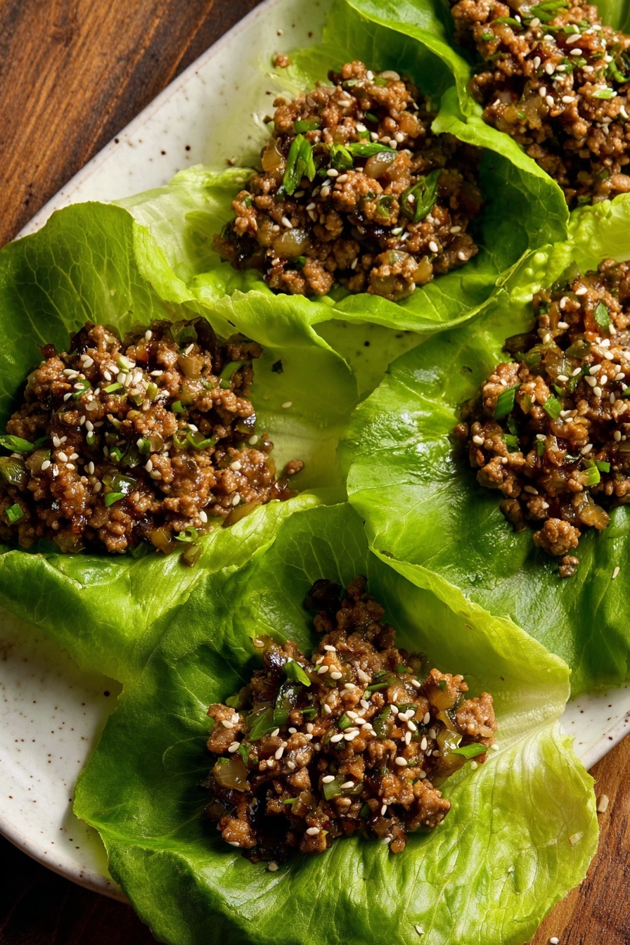 Tofu Lettuce Wraps, healthy tofu wraps, quick vegetarian dinner, crunchy tofu lettuce wraps, easy Asian lettuce wraps - The image shows five large green lettuce leaves arranged on a white plate with a wood surface in the background. Each leaf holds a serving of cooked ground meat mixed with small diced vegetables, with a brown color and a slightly moist texture. The meat mixture is sprinkled with white sesame seeds and small bits of green herbs. The lettuce leaves are fresh with visible veins and crinkled edges, creating a bright green contrast to the darker meat filling. Photo taken with an iphone --ar 2:3 --v 7