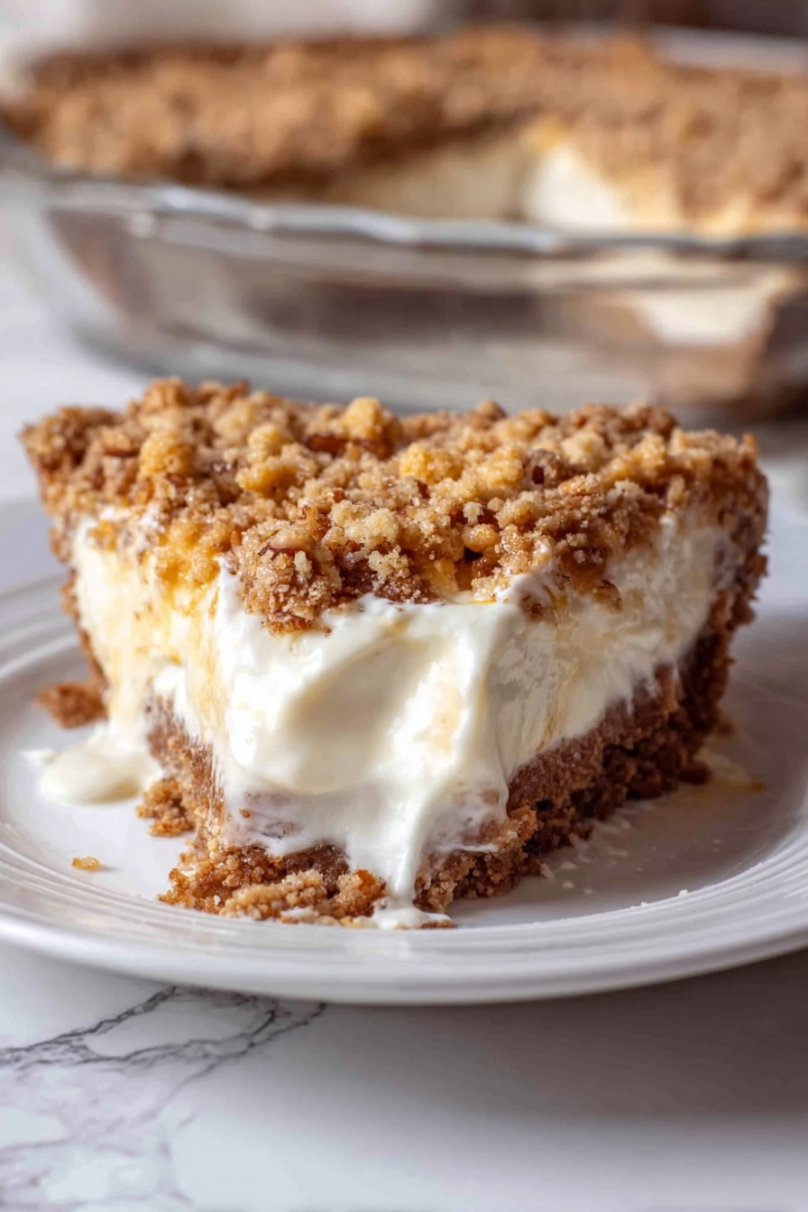 Angel Pecan Pie, pecan pie dessert, fluffy pecan pie, elegant pecan pie, easy holiday pies - A close-up of a slice of pie on a white plate with a white marbled surface underneath. The pie has three visible layers: a crumbly brown crust at the bottom, a thick creamy white filling in the middle that is slightly melting and spreading on the plate, and a chunky golden brown crumb topping with some small nut bits on top. In the background, part of a clear glass dish shows the rest of the pie with the same three layers. The photo taken with an iphone --ar 2:3 --v 7