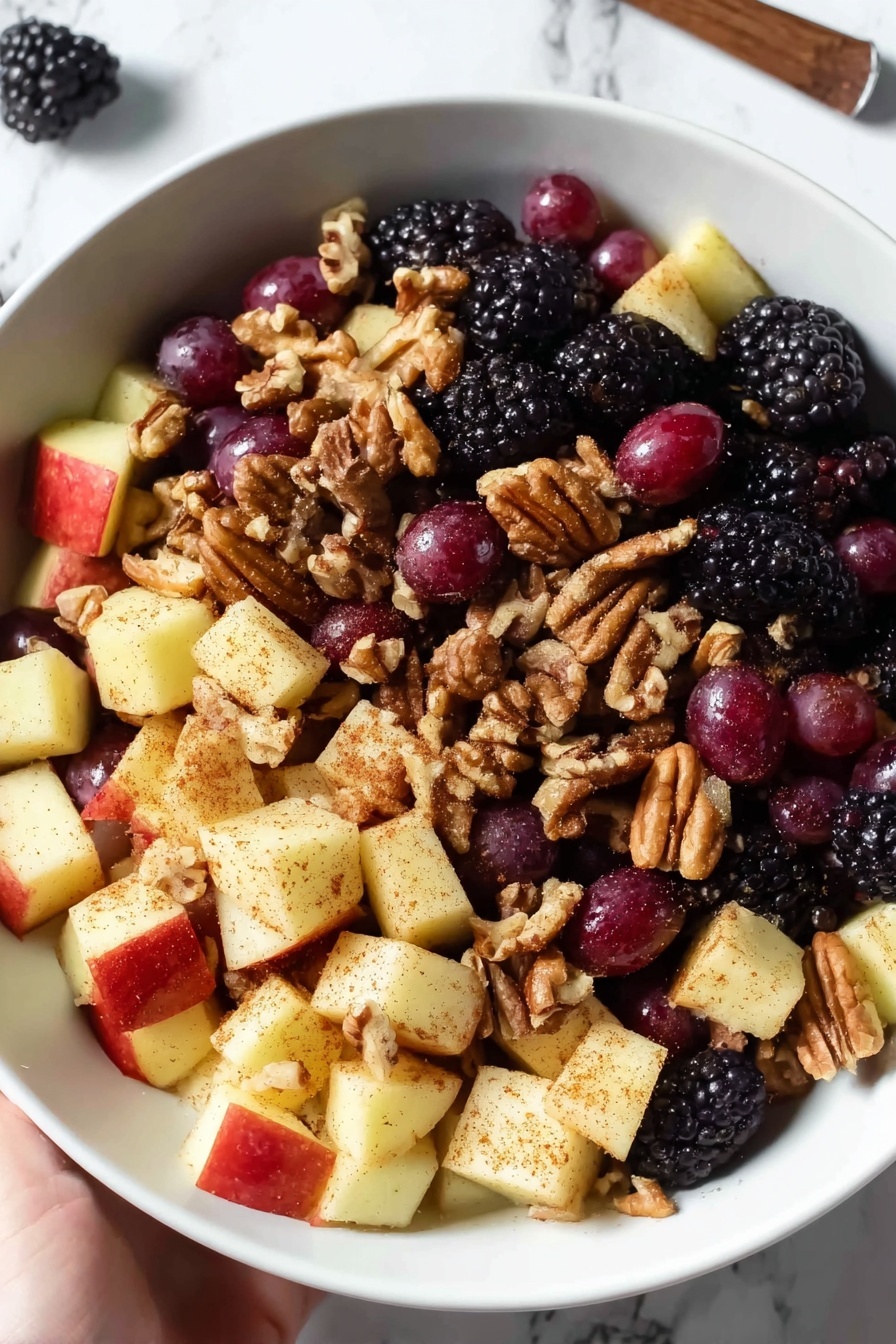 Fall Fruit Salad with Maple Cinnamon Dressing, fall fruit salad recipes, cozy fall fruit salad, healthy fall fruit salad, maple cinnamon dressing recipe - A white bowl is filled with a fruit salad that has three main layers: the bottom layer is made of blackberries, which are bumpy and dark purple-black; the middle layer has round red grapes with smooth, shiny skin; and the top layer contains apple chunks that are pale yellow with a dusting of brown cinnamon powder. Scattered throughout the salad are light brown pecans adding a rough texture. The bowl sits on a white marbled surface, and two cinnamon sticks lie nearby. photo taken with an iphone --ar 2:3 --v 7