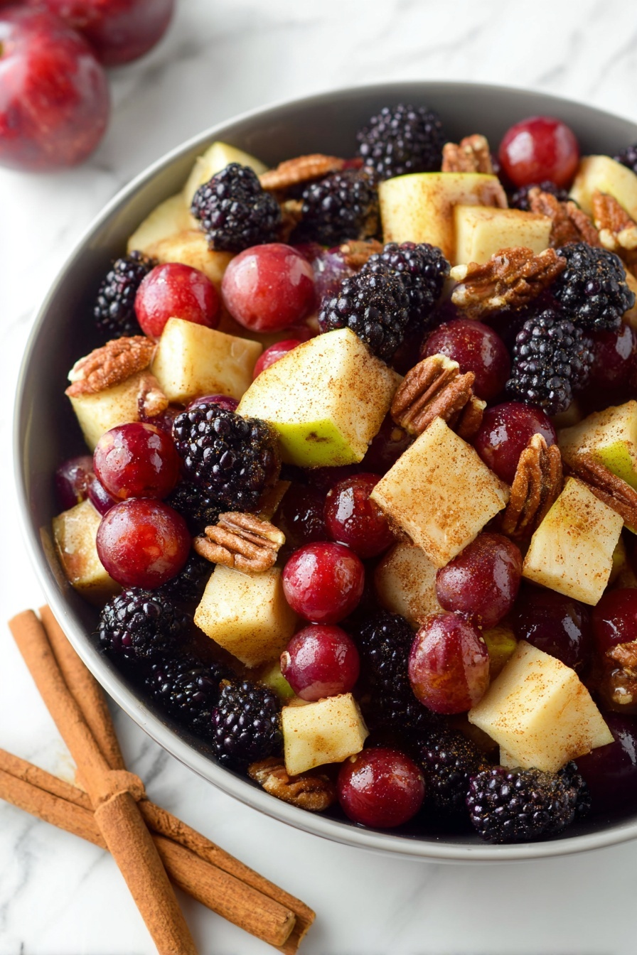 Fall Fruit Salad with Maple Cinnamon Dressing, fall fruit salad recipes, cozy fall fruit salad, healthy fall fruit salad, maple cinnamon dressing recipe - A white bowl filled with a colorful fruit and nut salad, showing three layers of ingredients. The bottom layer is pieces of pale yellow apple with red skin, cut into small cubes. The middle layer features deep red grapes and dark purple-black blackberries scattered evenly across. The top layer includes crunchy brown walnuts and pecans mixed throughout. The fruit pieces have a light dusting of cinnamon or spice, giving a slight matte texture. The bowl is placed on a white marbled surface, and a woman's hand is visible holding the bowl's edge. photo taken with an iphone --ar 2:3 --v 7