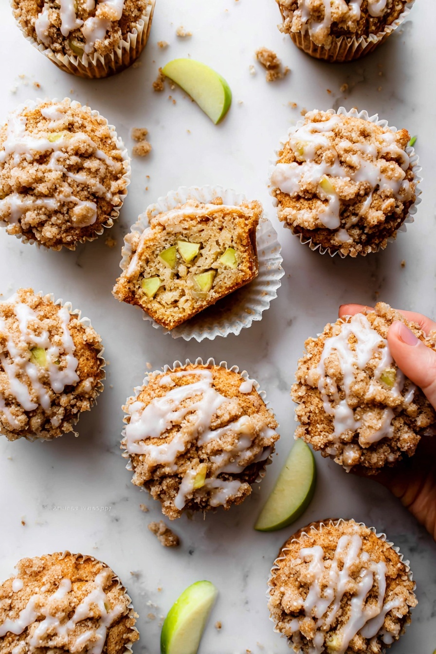 Apple Cinnamon Crumb Muffins, apple cinnamon muffins, easy apple muffins, homemade apple muffins, fall breakfast muffins - The image shows nine apple crumb muffins arranged on a white marbled surface. Each muffin has three clear layers: a bottom baked golden-brown cake layer, a middle layer with visible light green apple chunks inside the muffin, and a top crumbly light brown streusel topping with a slight sprinkle of white glaze drizzled unevenly over it. One muffin is open, showing the inside texture and apple pieces, sitting in a white paper liner with a woman's hand about to pick it up. Small crumbs and a few green apple slices are scattered around, adding detail to the scene. Photo taken with an iphone --ar 2:3 --v 7