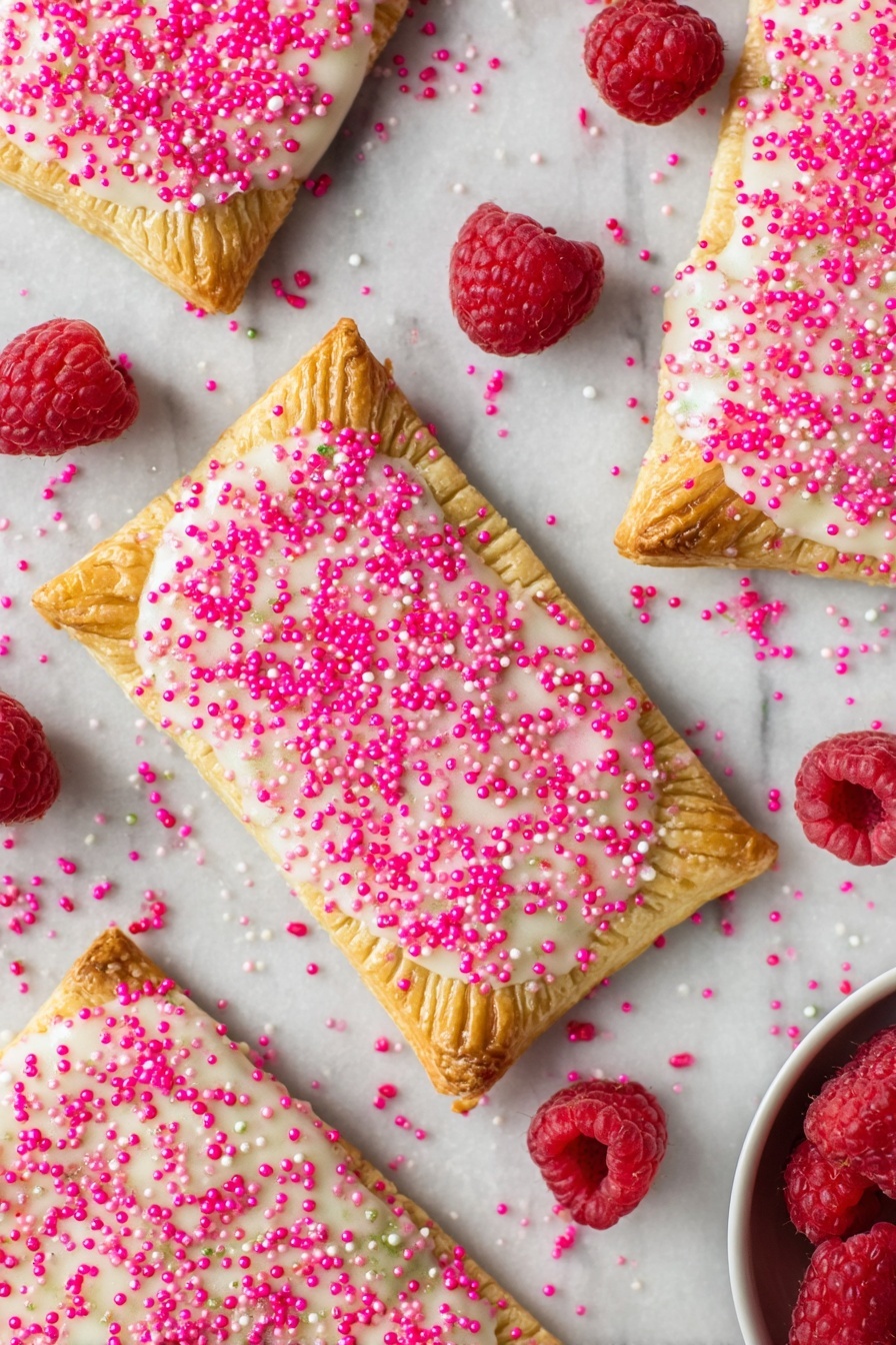 Homemade Raspberry Pop Tarts, raspberry pop tarts, homemade breakfast pastries, flaky pastry recipes, raspberry jam-filled pastries - Several rectangular toasted pastries spread over a white marbled surface, each with a golden-brown crust stamped with fork marks along the edges; the top layer is coated in a slightly shiny white icing sprinkled generously with bright pink sugar crystals, creating a textured look. Scattered around the pastries are fresh, plump red raspberries adding a pop of natural color. In the bottom right corner, a small portion of a white bowl is visible, holding more raspberries. photo taken with an iphone --ar 2:3 --v 7