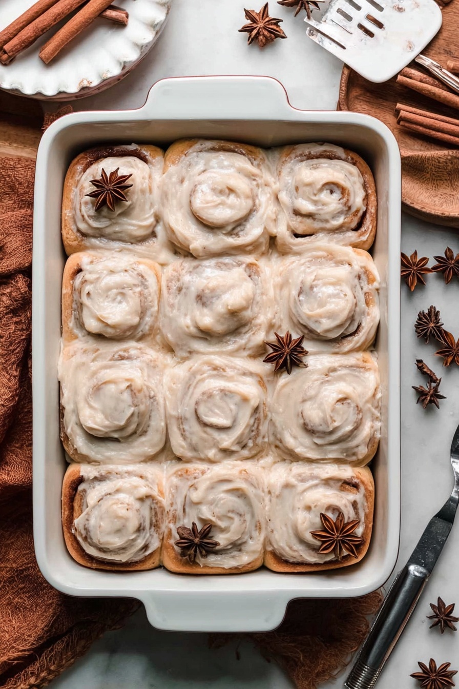 Chai Cinnamon Rolls with Caramel Sauce, chai cinnamon roll recipe, caramel chai rolls, weekend breakfast comfort, cozy chai dessert - A white rectangular baking dish filled with twelve cinnamon rolls topped with a thick, creamy light brown frosting, each roll showing distinct swirls and a soft texture. On top of the cinnamon rolls, there are a few whole star anise pieces scattered. The baking dish is placed on a white marbled surface with cinnamon sticks and star anise nearby, creating a cozy and warm atmosphere. A white scalloped plate and a metal spatula are partially visible at the edges. Photo taken with an iphone --ar 2:3 --v 7