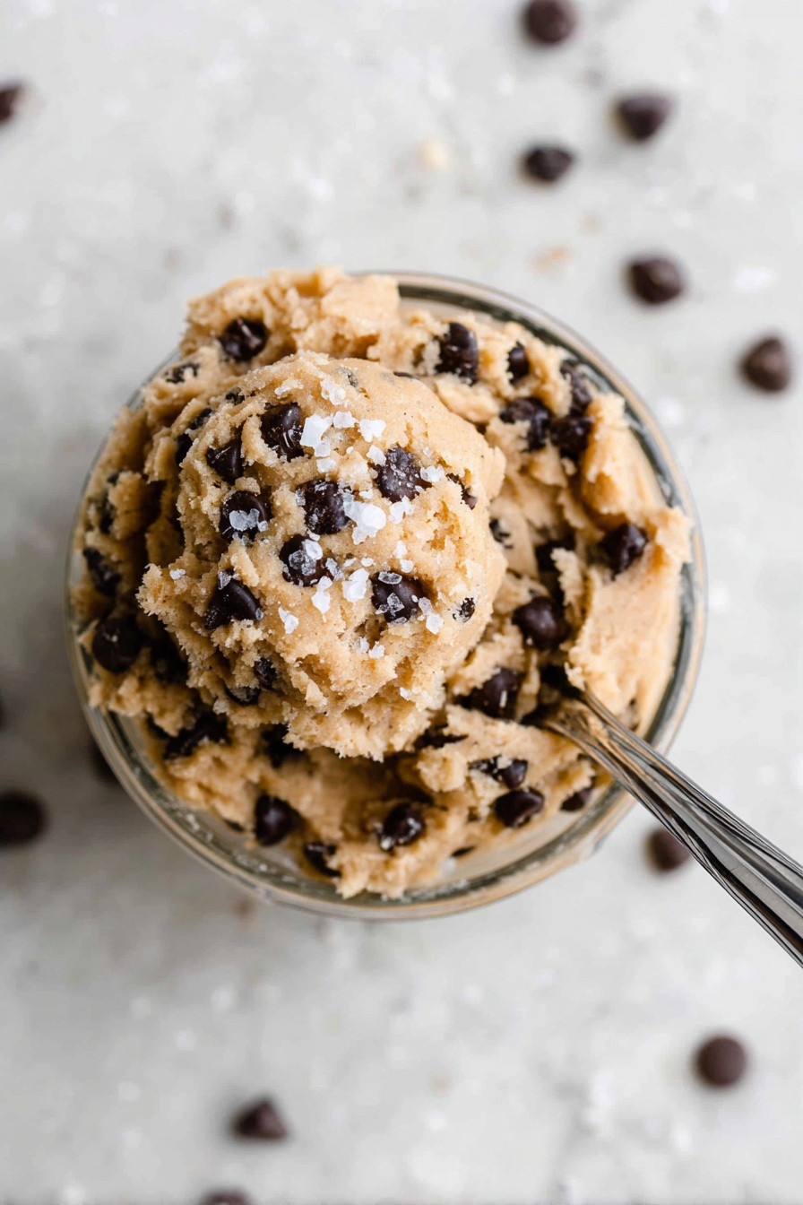Healthy Protein Cookie Dough Bites, healthy snack recipes, no-bake protein snacks, quick healthy treats, protein powder cookie bites - In a small clear glass bowl filled with one layer of light brown, soft cookie dough mixed with many small dark chocolate chips all over, the dough is slightly scooped at the top with a few coarse white salt flakes sprinkled on it. A shiny silver spoon is stuck into the dough from the right side, angled toward the top right corner, resting inside the bowl. The bowl sits on a surface with a white marbled texture, and some chocolate chips are scattered around it. The photo is taken with an iphone --ar 2:3 --v 7