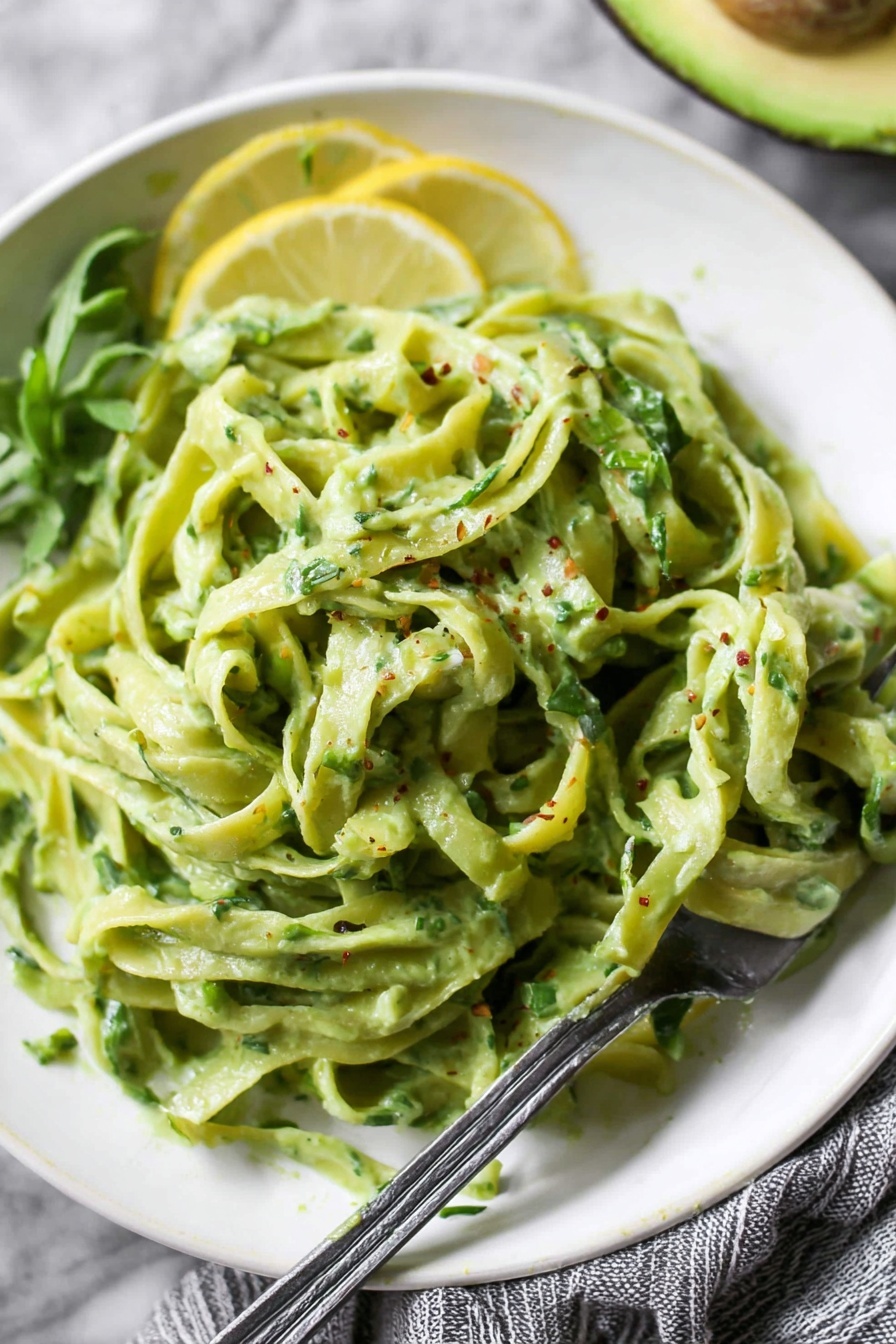 Avocado Lemon Pesto, healthy avocado pesto, quick vegan pesto, creamy lemon pesto, nut-free pesto - A white plate full of green pasta coated in a creamy sauce with hints of herbs, with thin green pasta ribbons layered loosely and a few small pieces of leafy herbs mixed in. On top left side of the plate, three thin lemon slices are placed for garnish. A silver fork is dug into the pasta from the right edge of the plate. In the background, there is a halved avocado with bright green flesh and a sprig of greenery on a white marbled surface. A white and gray striped cloth is partially visible on the right side. Photo taken with an iphone --ar 2:3 --v 7