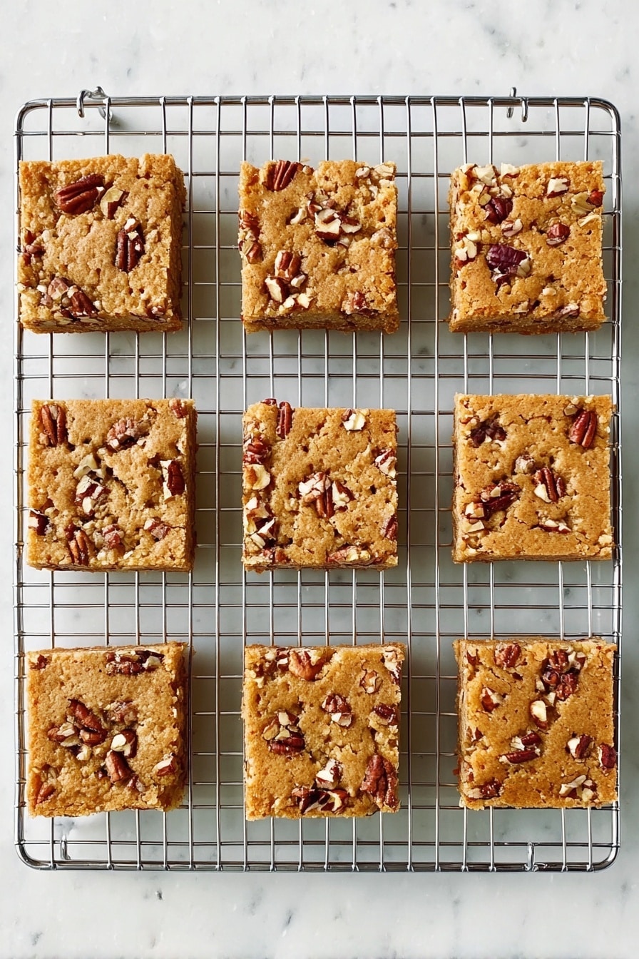 Maple Pecan Blondies, maple pecan blondies recipe, easy blondies with pecans, comforting fall desserts, homemade blondie bars - Nine square pieces of light brown nut bars sit on a silver cooling rack. Each bar has small, uneven chunks of pecans scattered on top, creating a rough texture. The bars are evenly spaced in three rows and three columns, showing a soft, baked surface. The background is a white marbled texture. Photo taken with an iphone --ar 2:3 --v 7