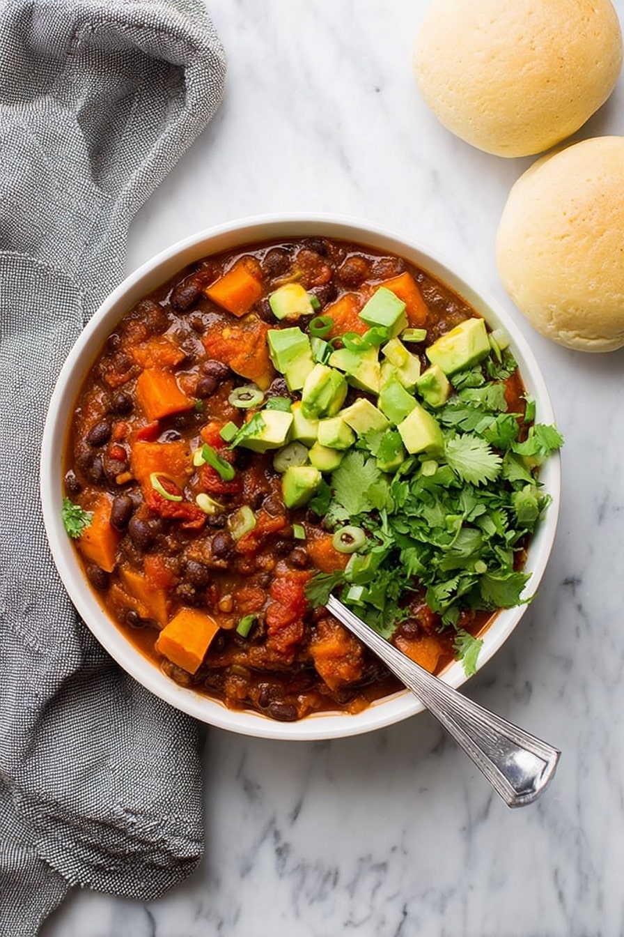 Sweet Potato Black Bean Chili, hearty vegetarian chili, easy healthy chili, cozy vegan chili recipe, nourishing plant-based chili - A white bowl filled with three main layers: the base is a thick stew of dark brown beans mixed with deep orange chunks of sweet potato and red tomato pieces in a reddish-brown sauce, topped on one side with bright green chopped avocado cubes and sprinkled with small rings of green onion, and on the other side with fresh green cilantro leaves. A silver spoon rests inside the bowl at the right side. The bowl sits on a white marbled surface next to two light beige round bread rolls and a loosely folded gray cloth. Photo taken with an iphone --ar 2:3 --v 7