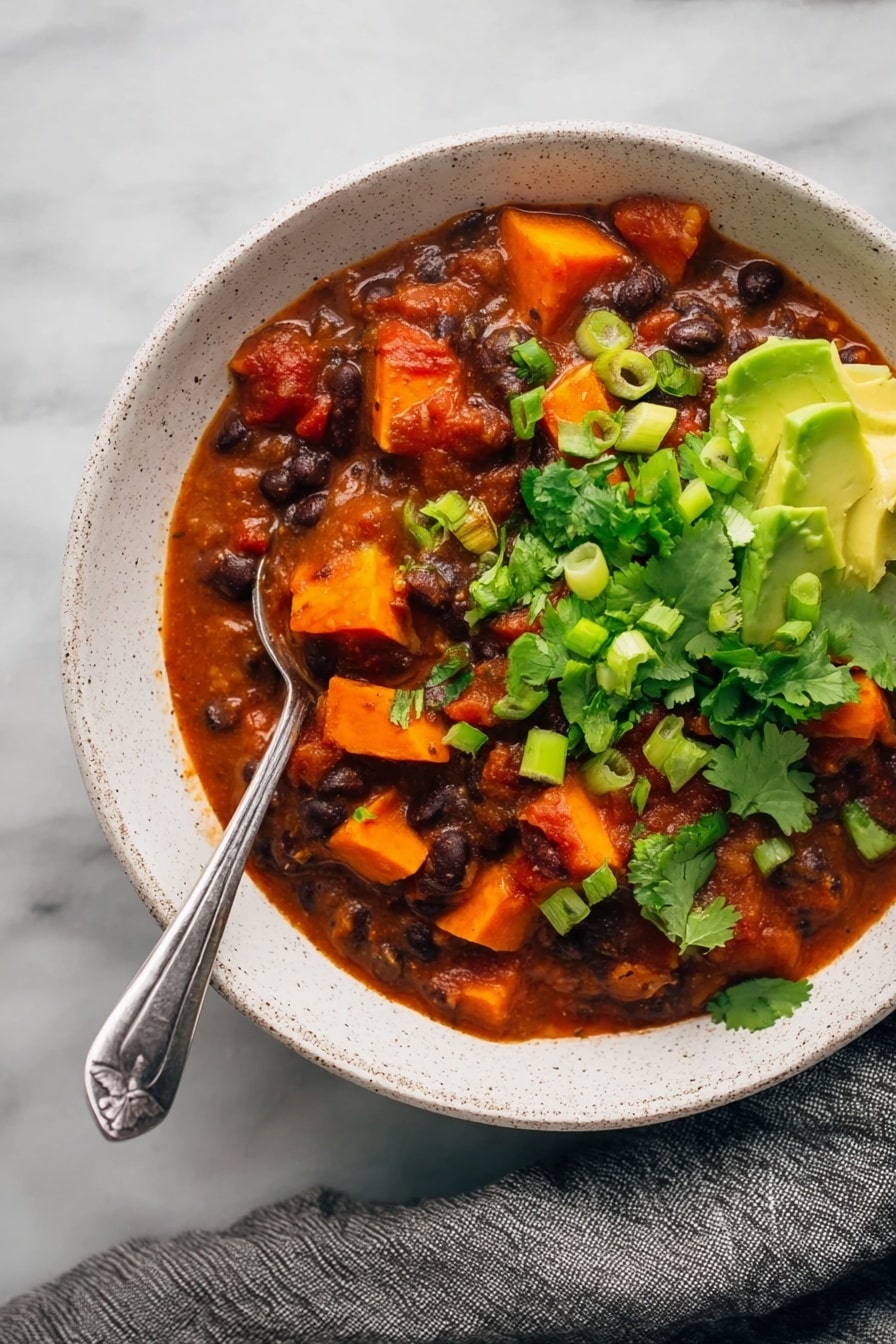 Sweet Potato Black Bean Chili, hearty vegetarian chili, easy healthy chili, cozy vegan chili recipe, nourishing plant-based chili - The image shows a white speckled bowl filled with a thick stew made of dark black beans and bright orange chunks of sweet potato in a rich red tomato sauce. The stew is garnished with vibrant fresh green cilantro leaves and sliced green onions, while creamy green avocado pieces sit on one side near the bowl’s edge. A silver spoon is placed in the stew with its handle resting on the edge of the bowl. The bowl is set on a white marbled surface with a gray cloth draped nearby. photo taken with an iphone --ar 2:3 --v 7