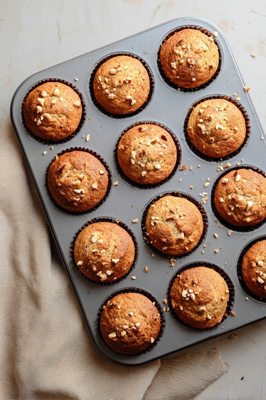 Quick & Easy Vegan Banana Muffins, vegan banana muffins, healthy banana muffin recipe, plant-based banana muffins, quick vegan breakfast muffins - A grey muffin tray holds twelve freshly baked muffins, each with a golden-brown top that is slightly rounded and textured with small cracks. Light tan chopped nuts are scattered on the surface of each muffin, adding a crunchy detail. The muffins sit in dark brown paper liners that are barely visible around the edges. The tray rests on a white marbled texture surface with a soft beige cloth partially visible on the left side. The light gently highlights the warm tones and textures of the muffins, making them look soft and fresh. photo taken with an iphone --ar 2:3 --v 7