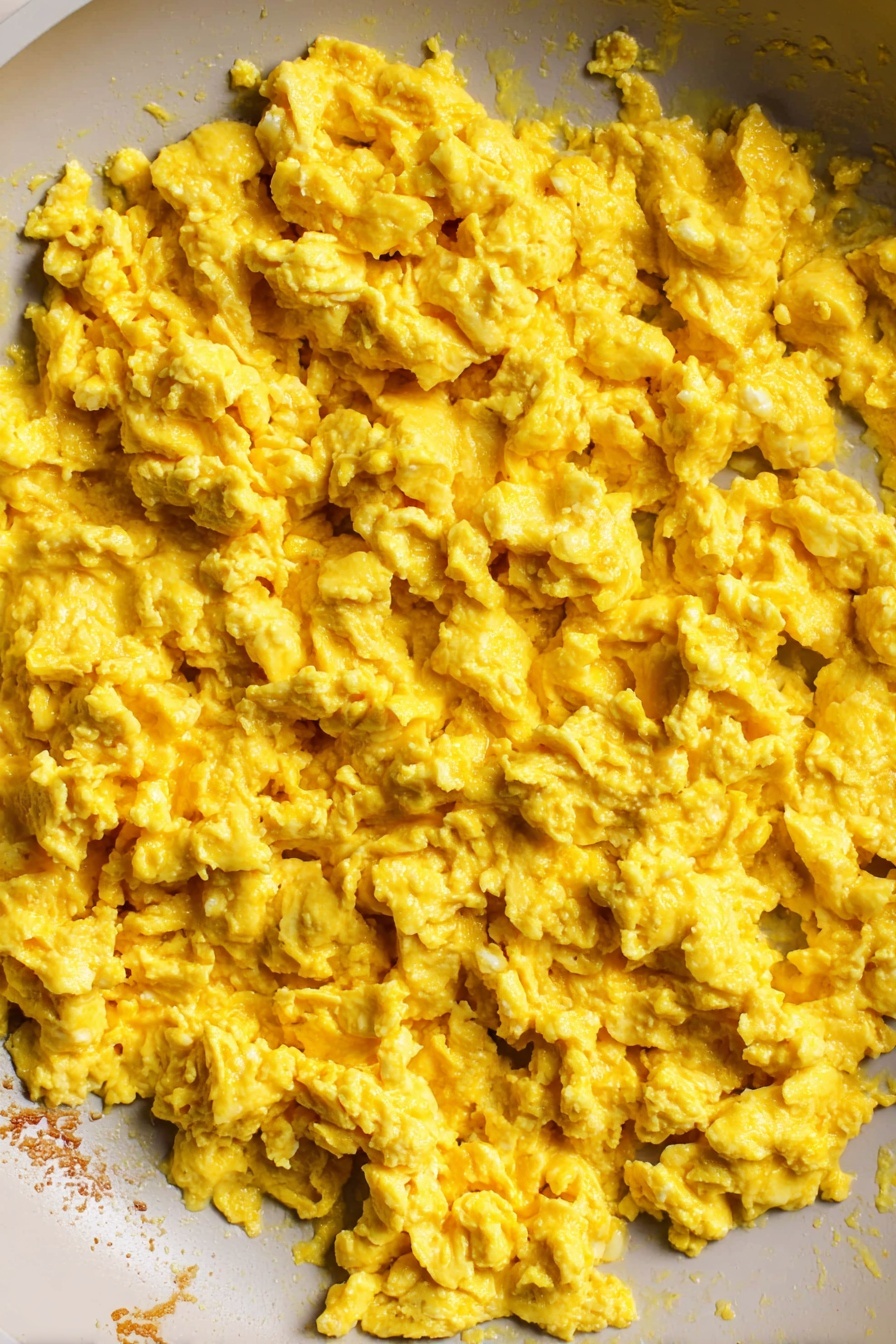 The Best Tofu Scramble, tofu scramble recipe, plant-based breakfast, vegan breakfast ideas, healthy tofu recipes - The image shows a close-up view inside a white pan filled with scrambled eggs. The cooked eggs have a soft, slightly chunky texture with a bright yellow color spread evenly across the pan. The edge of the pan reveals some lightly browned bits where the eggs touched the surface. The background is a white marbled texture. photo taken with an iphone --ar 2:3 --v 7