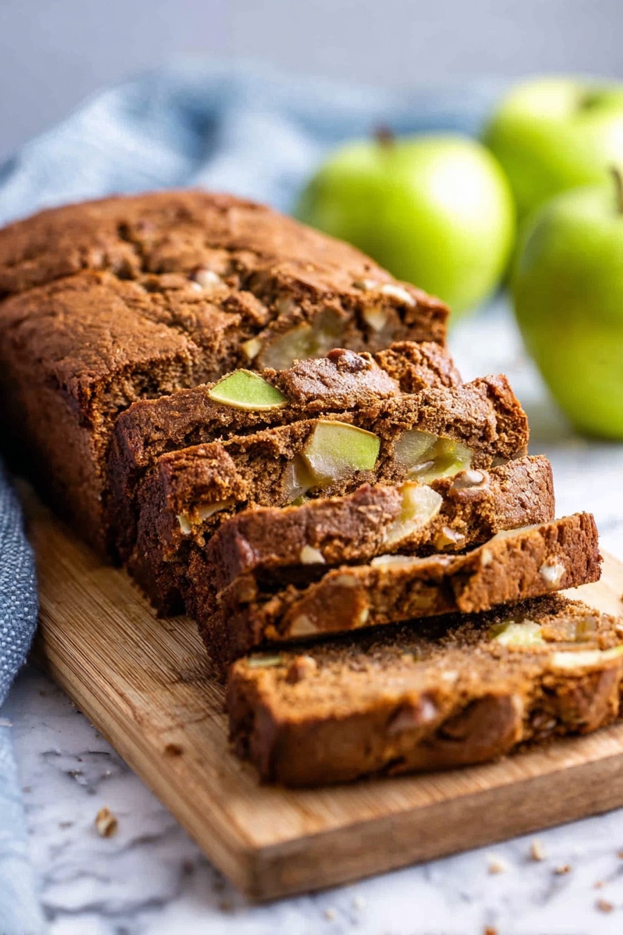 Moist Cinnamon Apple Bread, cinnamon apple bread, fall apple bread, cozy apple dessert, easy apple bread - A loaf of brown apple bread with a soft, cracked crust sits on a wooden board placed on a white marbled surface. The bread is sliced into thick pieces, revealing a moist inside with chunks of green apple and nuts scattered throughout. The background has a few whole green apples and a light blue kitchen towel, slightly blurred to keep the focus on the bread. photo taken with an iphone --ar 2:3 --v 7