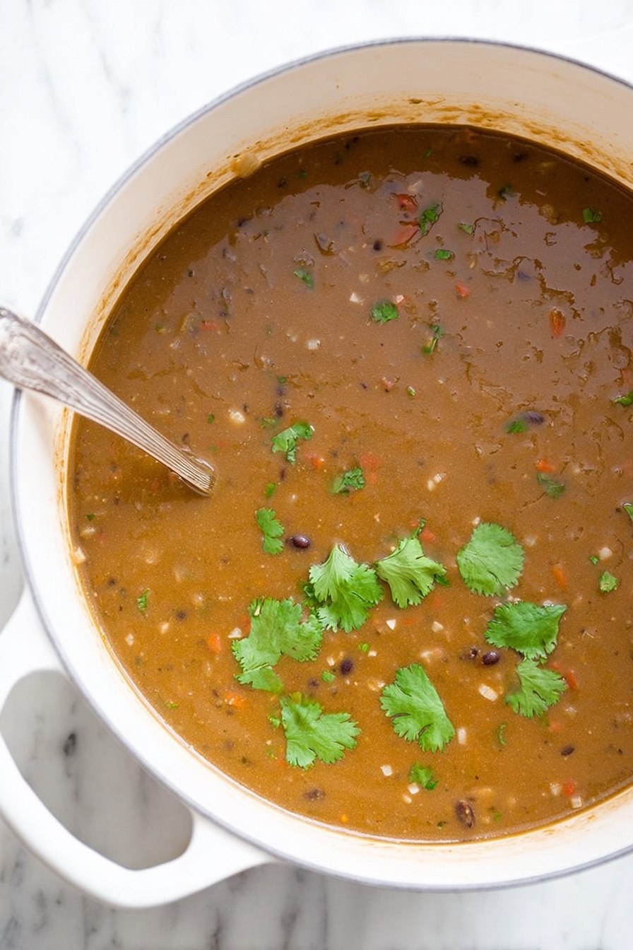 Vegan Black Bean Soup, vegan black bean soup savory dish, hearty vegan soup recipe, plant-based black bean soup, easy vegan soup ideas - A large white pot filled with thick brown soup that has a slightly grainy texture, small visible pieces of vegetables, and beans mixed throughout. Several fresh green cilantro leaves float on top, adding a touch of bright green color. A silver spoon is partially dipped into the soup on the left side of the pot. The pot is placed on a white marbled surface. Photo taken with an iphone --ar 2:3 --v 7