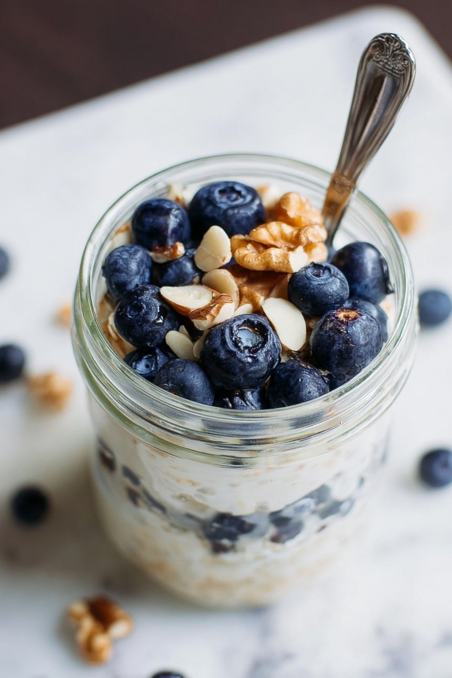 Simple Vegan Overnight Oats, vegan breakfast ideas, healthy vegan breakfast, easy vegan oatmeal, cruelty-free breakfast recipes - A clear glass jar filled with three layers: the bottom layer is fresh blueberries, the middle layer is creamy white porridge or yogurt with a smooth texture, and the top layer has more blueberries and a few light brown walnut pieces scattered on top. A silver spoon is placed in the jar, partly inside the food. The surface around the jar shows a white marbled texture with some scattered white nut pieces. Photo taken with an iphone --ar 2:3 --v 7