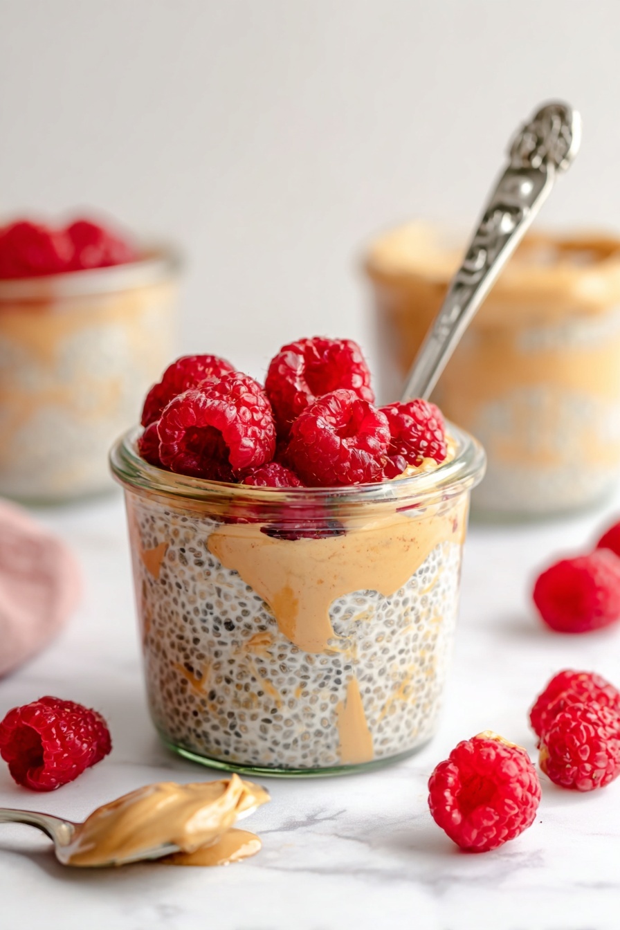 Peanut Butter Overnight Oats, healthy breakfast ideas, easy overnight oats, nutritious breakfast recipes, vegan overnight oats - A clear glass jar filled with three visible layers: the bottom layer is light beige chia seed pudding with black chia seeds mixed in, the middle layer shows swirls of creamy light brown peanut butter, and the top layer is a bright red cluster of fresh raspberries. A silver spoon with an ornate handle is placed inside the jar, leaning against the side with some pudding on it. Around the jar, scattered fresh raspberries add more red spots on a white marbled surface. In the background, more jars with similar contents and a silver spoon with a light brown spread are softly blurred. Photo taken with an iphone --ar 2:3 --v 7