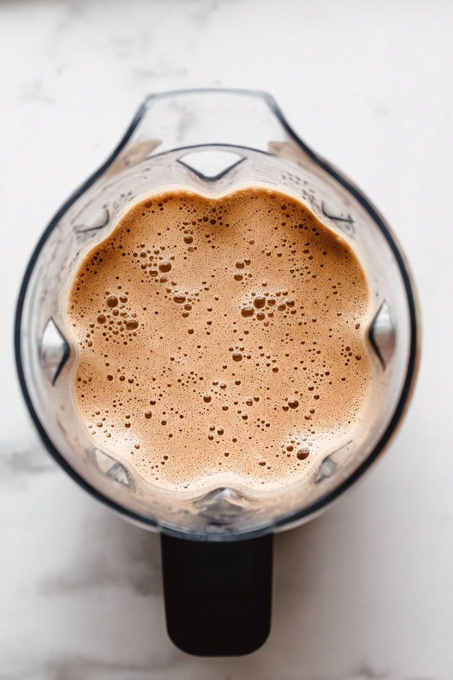 Vegan Chocolate Peanut Butter Protein Shake, healthy vegan protein shakes, plant-based chocolate peanut butter smoothie, quick vegan protein drinks, dairy-free chocolate peanut butter shake - The image shows a clear blender from above filled with a light brown, frothy mixture with many small bubbles on its surface. The blender has a black handle, and the background is a white marbled surface. photo taken with an iphone --ar 2:3 --v 7