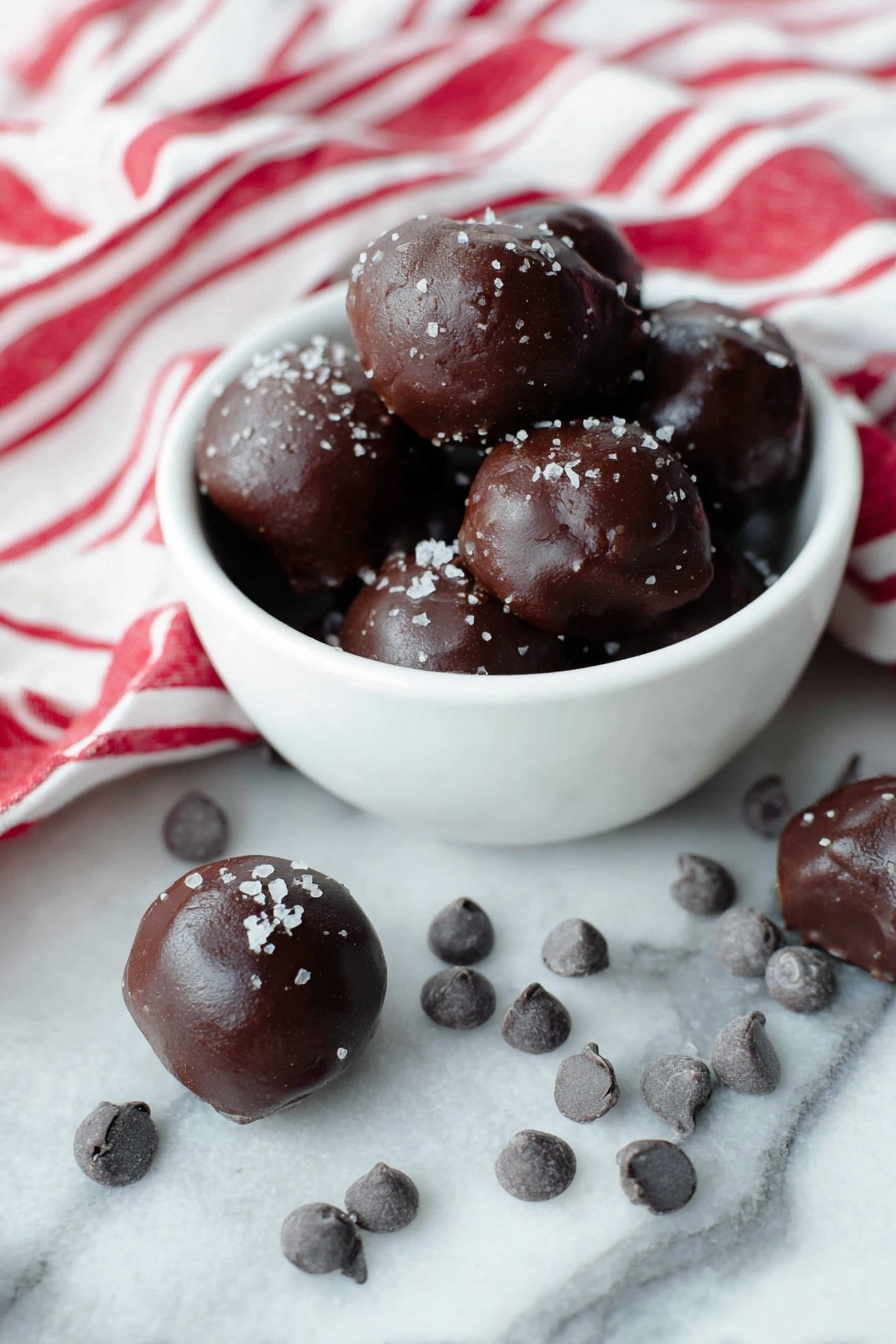 Healthy Cocoa Date Truffles, healthy chocolate treats, nutritious date balls, best guilt-free desserts, simple wholesome snacks - A white bowl filled with several dark brown chocolate balls that have a smooth, shiny texture and are sprinkled lightly with coarse salt flakes on top. Around the bowl, more chocolate balls sit on a white marbled surface, some resting near small clusters of dark chocolate chips. A white cloth with bold red stripes is casually draped to the back and right side of the bowl, adding a bright contrast. The scene is bright and clean, showing the rich dark color and glossy texture of the chocolates clearly. Photo taken with an iphone --ar 2:3 --v 7