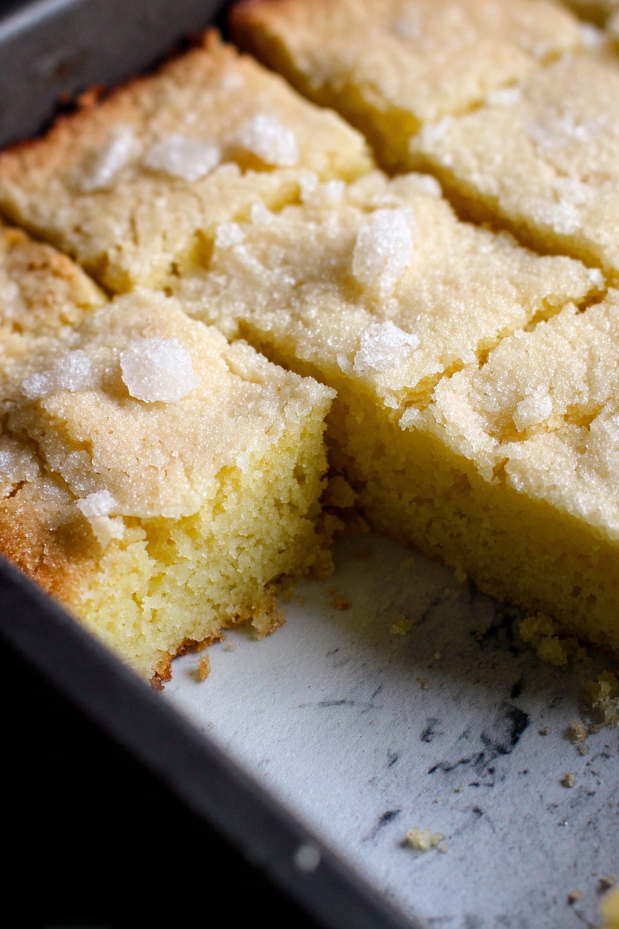 French Butter Cake, French Butter Cake recipe, buttery dessert, easy French cake, tender butter cake - A single-layer light yellow cake is shown inside a dark baking pan with a white marbled texture surface underneath. The top layer has a rough crumbly texture with sugar crystals scattered on top, and it is cut into square pieces with one piece missing, revealing a soft, dense inside that is smooth and slightly moist. The cake edges are slightly browned and cracked, contrasting with the pale yellow center. Photo taken with an iphone --ar 2:3 --v 7