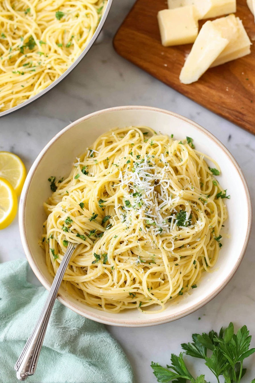 Lemon Garlic Pasta, Lemon Garlic Pasta Recipe, Easy Lemon Pasta, Zesty Garlic Pasta, Bright Lemon Pasta - A white bowl filled with light yellow spaghetti pasta mixed with small green herb bits, topped with fine white cheese shavings and fresh green parsley. On the side of the pasta are two lemon wedges. A silver fork rests inside the pasta on the left side. In the background, there is a white pan with more spaghetti and a wooden board with chunks of light yellow cheese and green parsley on a white marbled surface. A light green cloth is near the bottom left corner. Photo taken with an iphone --ar 2:3 --v 7