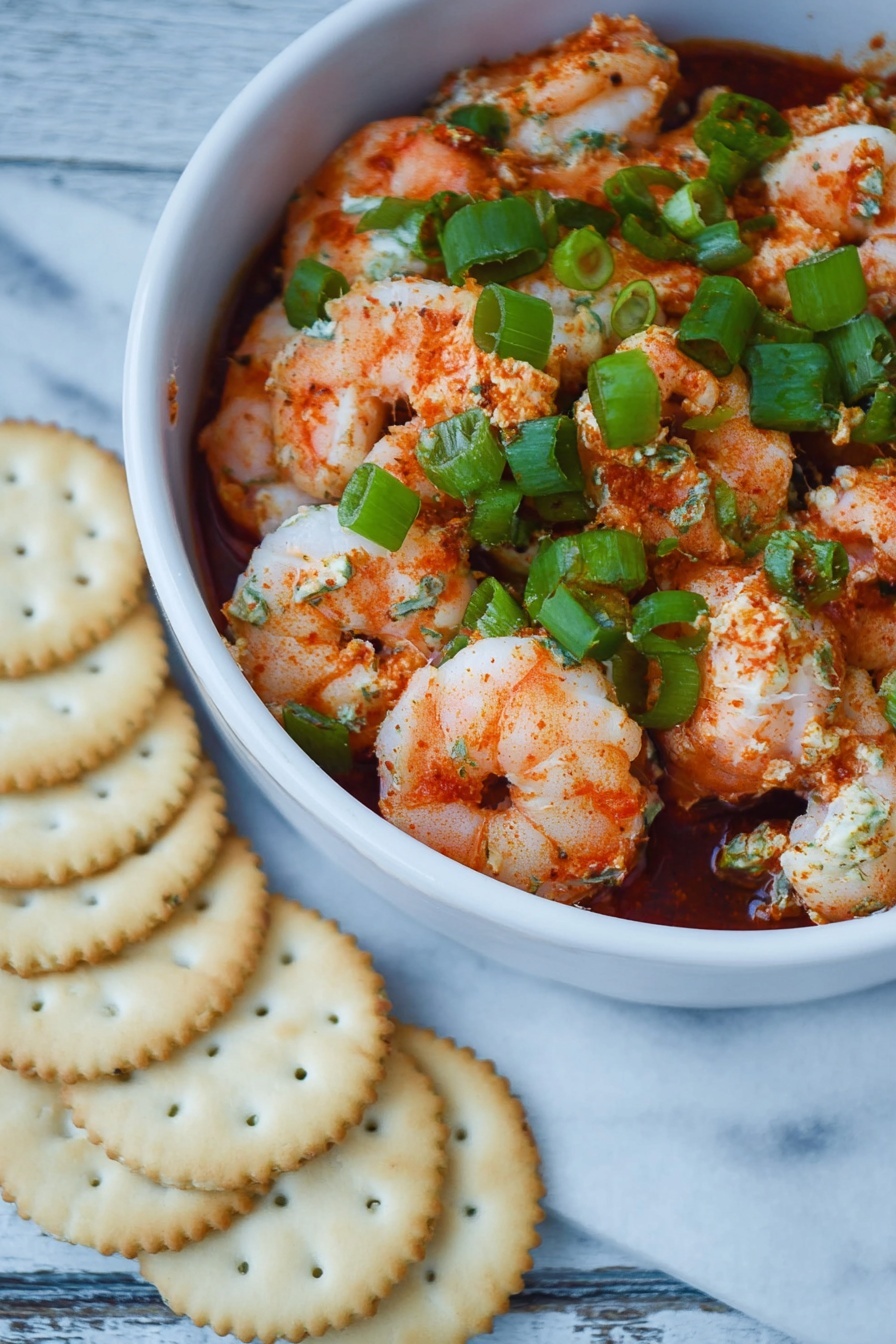 Shrimp Cocktail Dip, seafood appetizer recipes, easy party dips, creamy shrimp dip, quick appetizer ideas - The dish shows one white bowl filled with three visible layers: a bottom layer of dark red sauce, a middle layer of pink and white cooked shrimp coated lightly in red spices, and a top layer of bright green chopped scallions scattered evenly over the shrimp. The textures include soft, plump shrimp with some seasoning powder visible and fresh, crisp scallions. The bowl is on a surface of white marbled texture, next to a semi-circle of pale golden round crackers arranged closely in front of the bowl. photo taken with an iphone --ar 2:3 --v 7