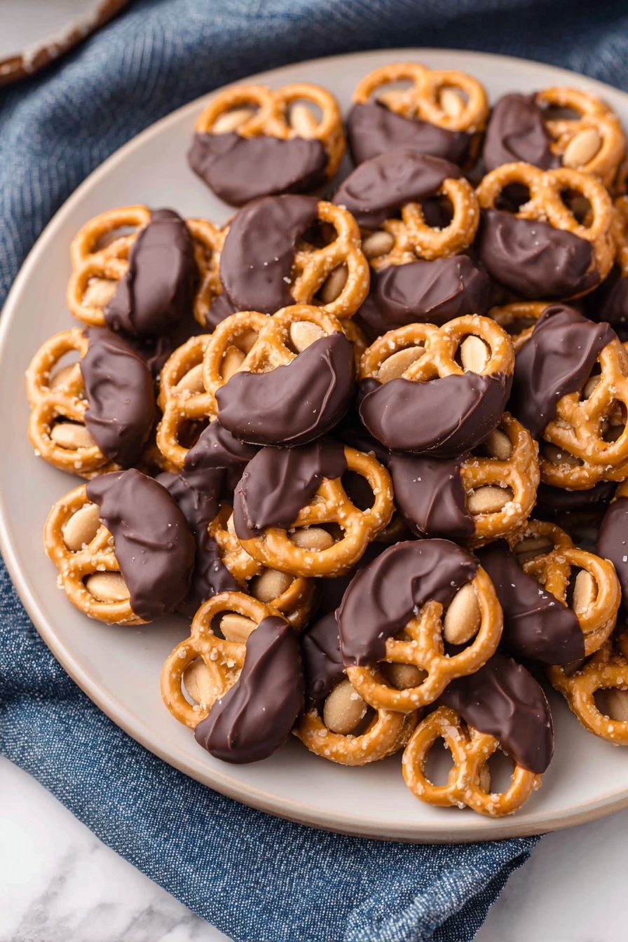Chocolate-Dipped Peanut Butter Pretzel Bites, sweet salty snack, no-bake treat, chocolate peanut butter bites, easy party snacks - A white plate filled with many small pretzels, each topped with a single peanut and half dipped in smooth dark chocolate, giving a mix of shiny black and golden tan colors. The pretzels have a crunchy texture with visible salt grains on the surface, while the peanuts underneath add a light brown soft contrast. The plate sits on a white marbled texture with a blue cloth nearby. photo taken with an iphone --ar 2:3 --v 7