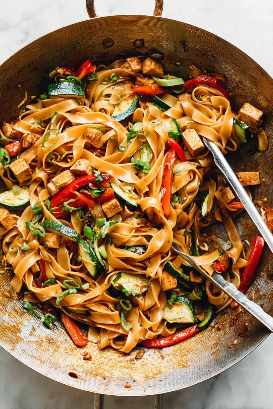 Drunken Noodles with Chicken and Basil, Thai stir-fry with chicken and basil, spicy chicken basil noodles, easy Thai drunken noodles, flavorful chicken stir-fry - The image shows a metal wok filled with a colorful stir-fry dish. The base layer consists of wide, flat noodles that are light brown and glossy. Mixed through the noodles are pieces of lightly browned tofu cubes, thin strips of green bell peppers, slices of red bell peppers, and small chunks of zucchini with their green skin visible. Scattered over the top are thinly sliced green onions and bits of chopped herbs adding dots of bright green. The noodles and vegetables appear shiny from a light sauce coating them evenly, and the wok has a warm, well-used appearance with some browning on the sides. A pair of metal tongs rests inside the wok, ready to serve. The setting has a clean, white marbled texture surface. photo taken with an iphone --ar 2:3 --v 7