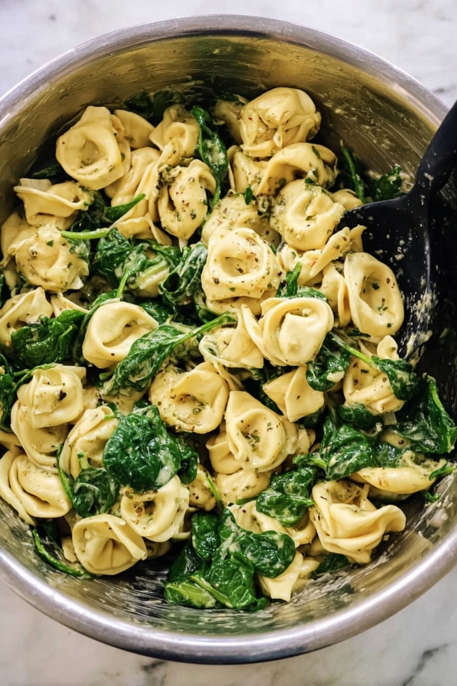 Pesto Tortellini, quick tortellini recipes, easy pasta dinners, cheesy tortellini with pesto, simple weeknight meals - This image shows a big metal bowl filled with cooked tortellini pasta mixed with bright green spinach leaves. The pasta is light yellow with a soft texture, and some pieces have dark green herb specks on them. The spinach looks fresh and slightly wilted, spread evenly throughout the pasta. There is a black spoon resting inside the bowl, ready to serve. The bowl sits on a white marbled surface, giving a clean and simple background. Photo taken with an iphone --ar 2:3 --v 7