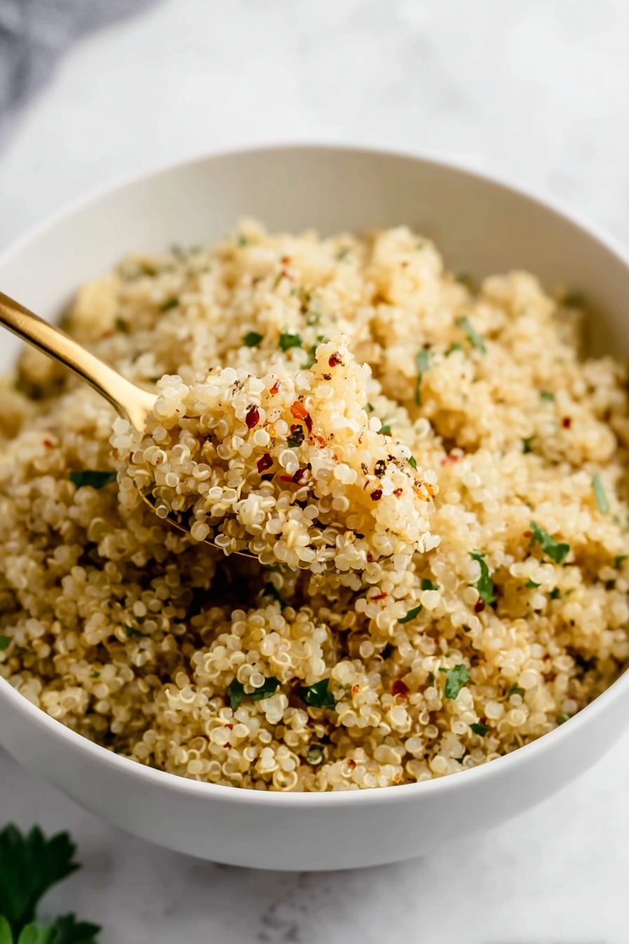 Garlic Butter Quinoa, flavorful quinoa side dish, easy garlic butter quinoa, quick quinoa recipes, best side dishes with quinoa - A close-up of a white bowl full of cooked quinoa, which is light beige and fluffy. The quinoa grains are mixed with small green herb pieces scattered evenly throughout and seasoning specks of black and red on top. A gold spoon scoops up some quinoa from the bowl, showing the grain’s soft, slightly textured surface. The bowl sits on a white marbled texture surface with some green leaves blurred in the background. photo taken with an iphone --ar 2:3 --v 7