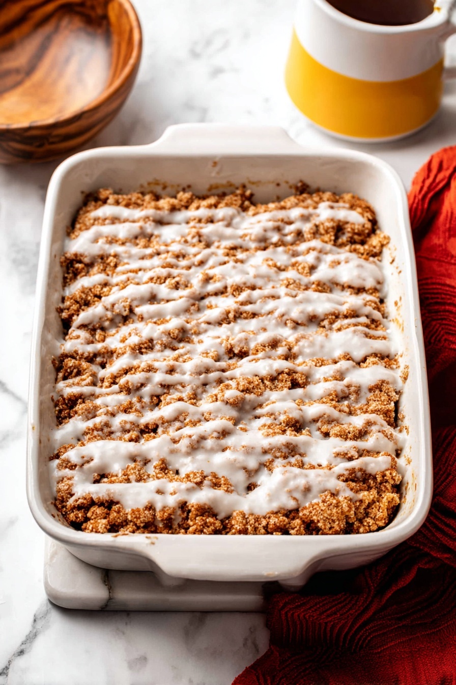 Pumpkin Crumb Coffee Cake, Pumpkin Crumb Coffee Cake recipe, Pumpkin spice coffee cake, Fall coffee cake with crumb topping, Pumpkin dessert with glaze - The image shows a white rectangular baking dish filled with a crumbly brown dessert layer, topped with a drizzle of white icing spread unevenly across the surface. The crumb layer has a soft, rough texture with patches of darker and lighter brown. The dish is placed on a white marbled surface, with part of a wooden bowl visible on the left and a yellow and white mug on the right. A red cloth lies partially beneath the baking dish at the bottom right corner. Photo taken with an iphone --ar 2:3 --v 7