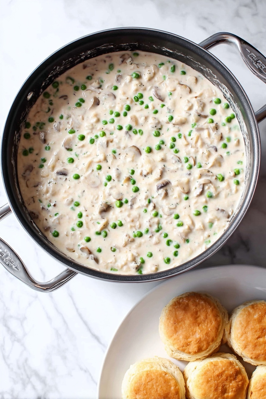 Creamed Chicken Over Biscuits, creamy chicken and biscuits, comforting chicken dinner, easy chicken recipes, quick dinner ideas - A round black pan filled with creamy white sauce that has small green peas and small light brown mushroom pieces mixed evenly throughout. The sauce looks thick and smooth. Next to the pan is a white plate holding about six golden brown biscuits stacked loosely. All is placed on a white marbled surface. photo taken with an iphone --ar 2:3 --v 7