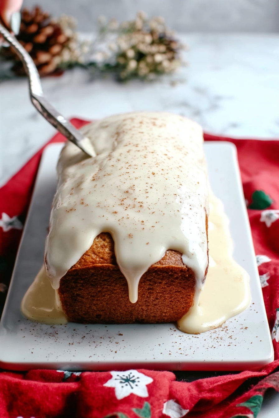 Eggnog Bread with Icing, holiday eggnog bread, festive bread recipes, Christmas flavored bread, easy eggnog dessert bread - A rectangular loaf cake sits on a white rectangular tray over a red cloth with white and blue flower patterns, placed on a white marbled surface. The cake has a golden-brown color and is covered on top with thick, creamy white icing that drips over the edges and pools around the base. The icing is lightly sprinkled with a fine brown powder, likely cinnamon or nutmeg. The scene includes dried flowers blurred in the background and a woman's hand holding a silver knife positioned above the cake. Photo taken with an iphone --ar 2:3 --v 7