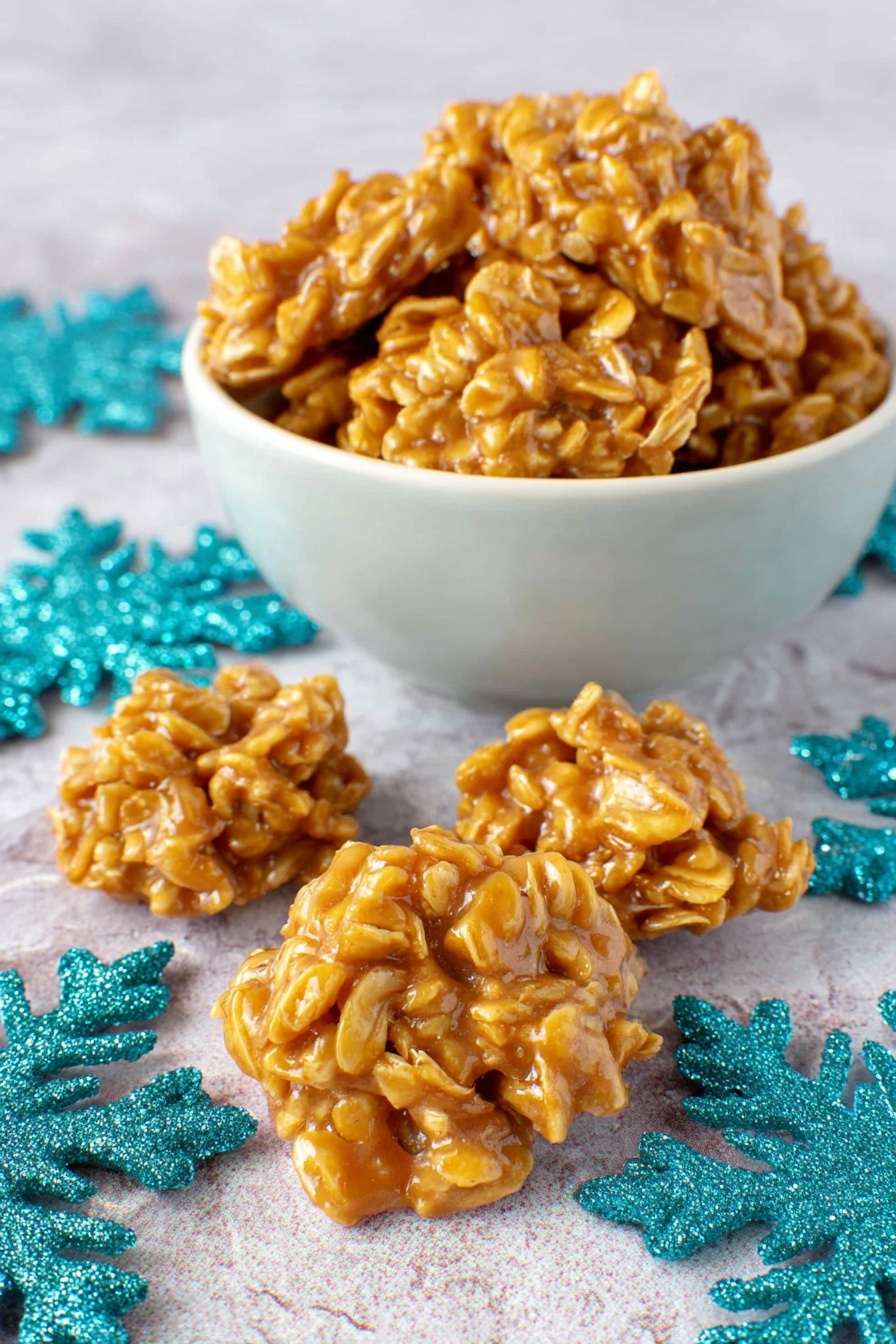 Peanut Butter Cornflake Candy, easy peanut butter candy, crunchy cornflake treats, homemade holiday candy, no-bake peanut butter snacks - The image shows a white bowl filled with golden brown no-bake cookie clusters that have a rough, lumpy texture with visible oats and a shiny glaze. In front of the bowl, there are four cookie clusters arranged on a white marbled surface, surrounded by blue and teal sparkly paper snowflakes for decoration. The clusters look slightly irregular in shape, each made of many overlapping layers of oats coated with a sticky caramel-colored syrup. photo taken with an iphone --ar 2:3 --v 7