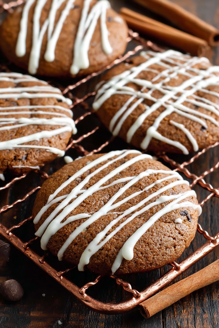 Soft Gingerbread Cookies, holiday gingerbread cookies, chewy gingerbread cookies, spiced gingerbread cookies, easy gingerbread cookies - The image shows four round brown cookies with a slightly cracked surface, placed on a copper cooling rack. Each cookie is decorated with thin, white icing drizzled randomly across the top, creating a light pattern. The cookies look soft and moist with dark spots indicating spices or chocolate chips. The background includes a dark wooden table with two cinnamon sticks placed on the right side. photo taken with an iphone --ar 2:3 --v 7