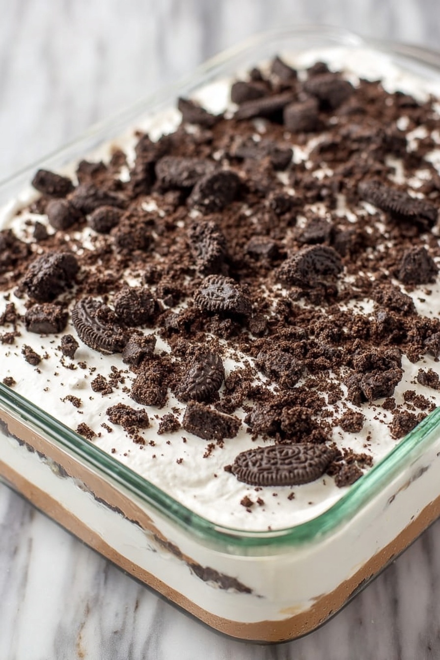 Red Velvet Poke Cake with Cheesecake Filling, Red Velvet Poke Cake, Cheesecake Poke Cake, Easy Red Velvet Cake Recipe, No-Bake Cheesecake Cake - A clear glass rectangular dish holds a layered dessert with three visible layers. The bottom layer is a light brown creamy base, covered by a thick middle layer of smooth white cream. The top layer is sprinkled generously with roughly crushed dark chocolate sandwich cookies, scattered evenly across the white cream. The dish sits on a white marbled surface. photo taken with an iphone --ar 2:3 --v 7